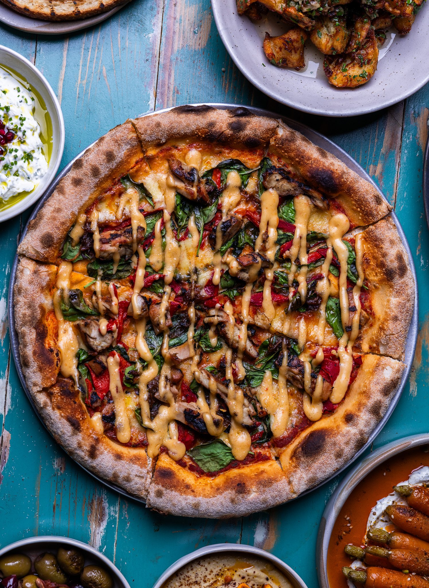 A pizza with cheese, spinach, red peppers, mushrooms, and drizzled sauce on top, surrounded by various bowls of side dishes.