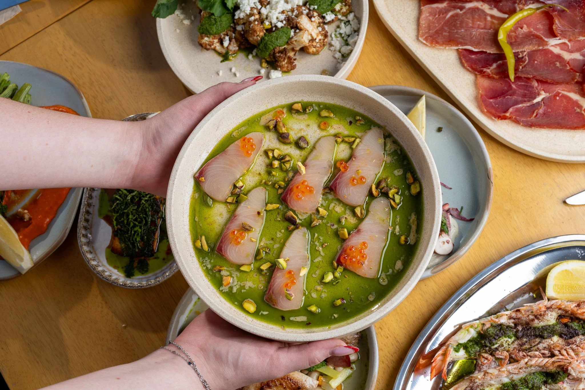 A person holding a bowl of sashimi topped with pistachios and salmon roe, surrounded by various dishes including cured meats, vegetables, and seafood on a wooden table.