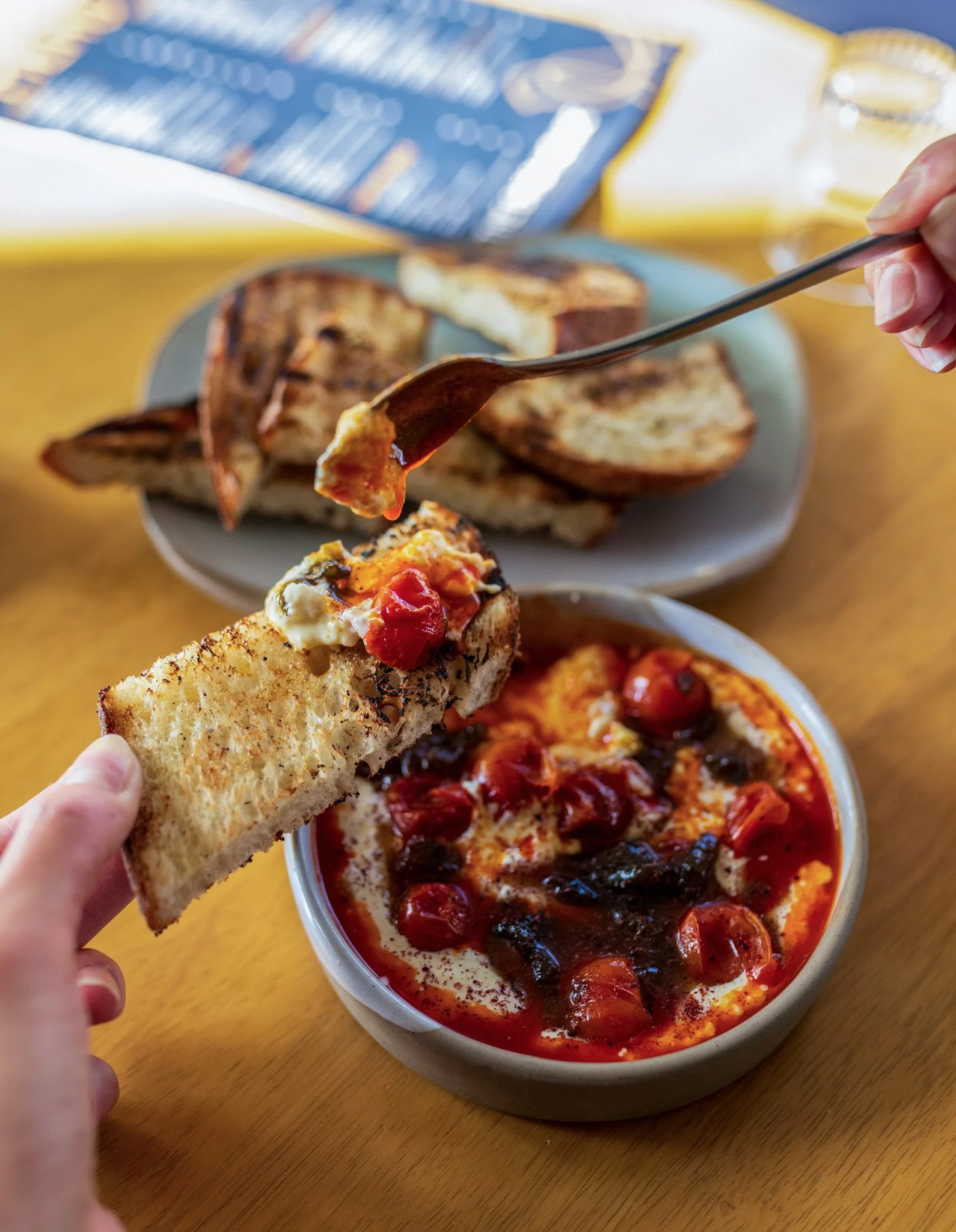 Person dipping toast into a bowl of chili with cherry tomatoes and black beans. Toasts are also on a plate in the background.