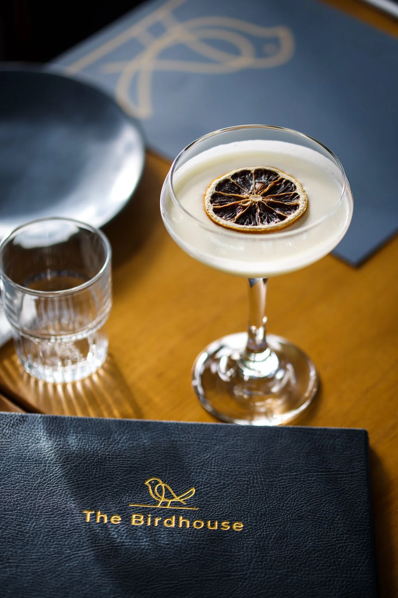 A cocktail glass with a lemon slice garnish on top, placed on a wooden table near a black menu or notebook with 'The Birdhouse' written on it, alongside a small glass of water, with a blue napkin and a silver plate in the background.