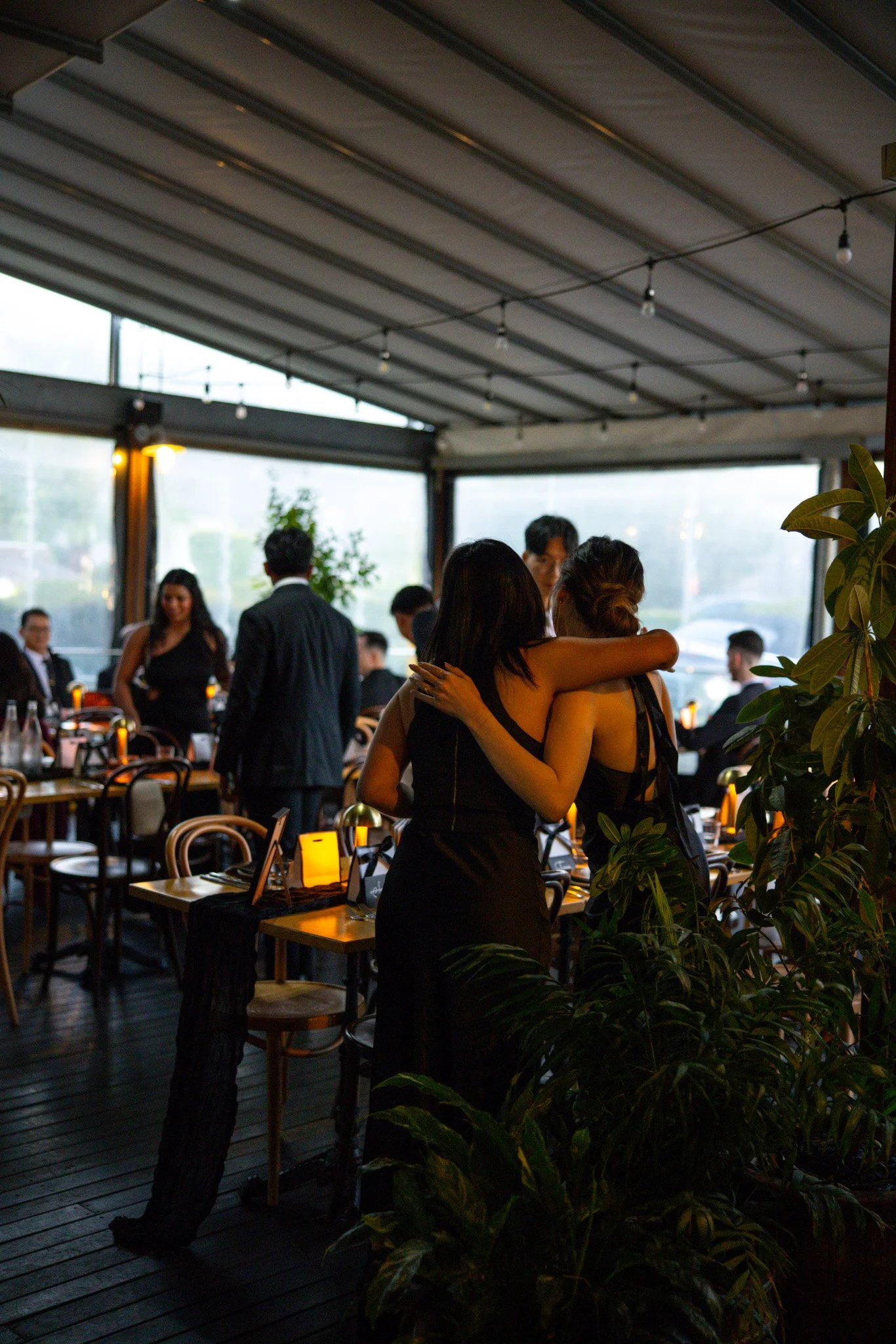 People talking and socializing inside a dimly lit restaurant or event space with tables, chairs, and warm lighting.