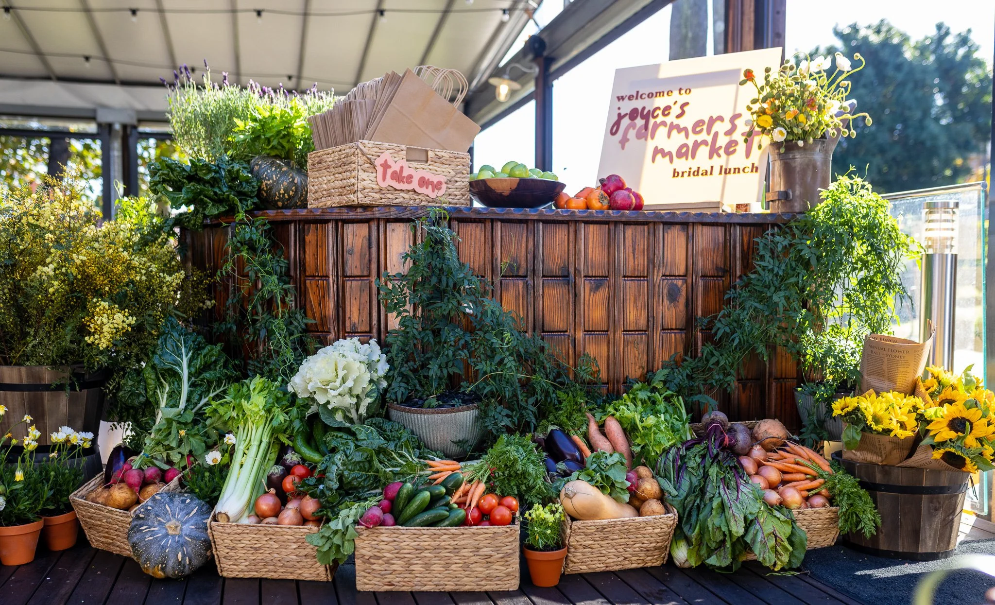 Market stand with various fresh vegetables and plants, including carrots, radishes, zucchini, and leafy greens, with signs and potted flowers.
