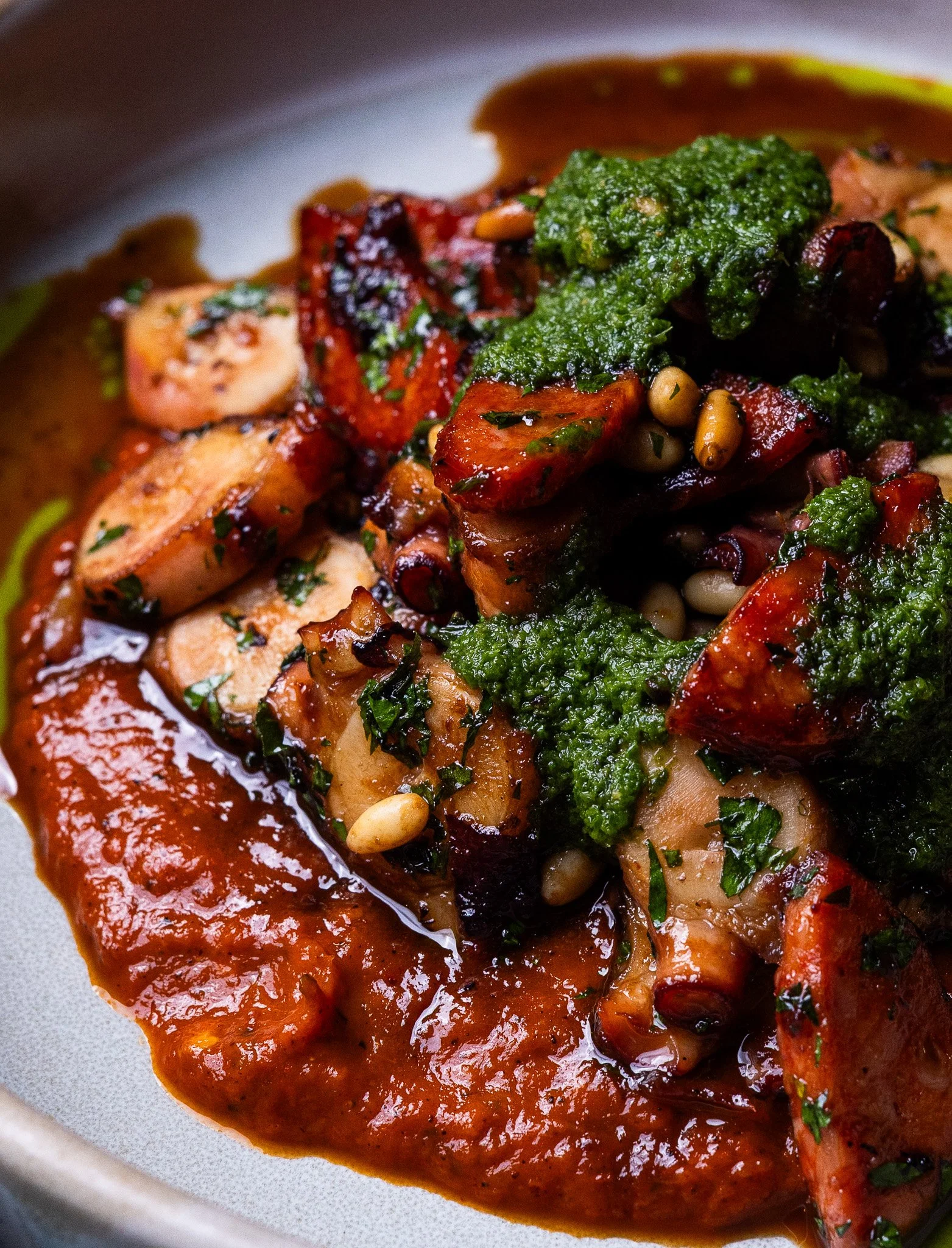 Close-up of a plated dish with grilled chicken, vegetables, pine nuts, and green sauce on top, served on a gray plate.