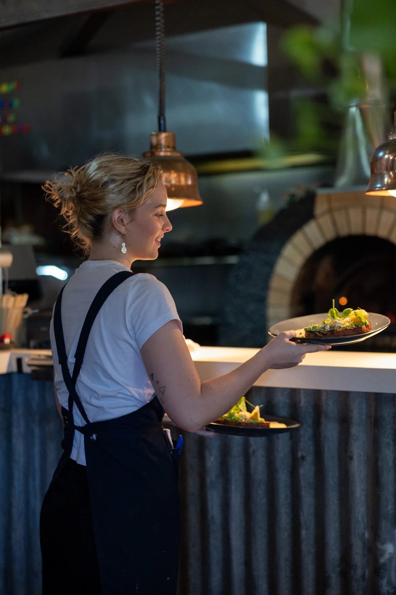 A woman with blonde hair tied up, wearing a white t-shirt and black apron, serving food in a restaurant kitchen. She is holding a plate with food and smiling.