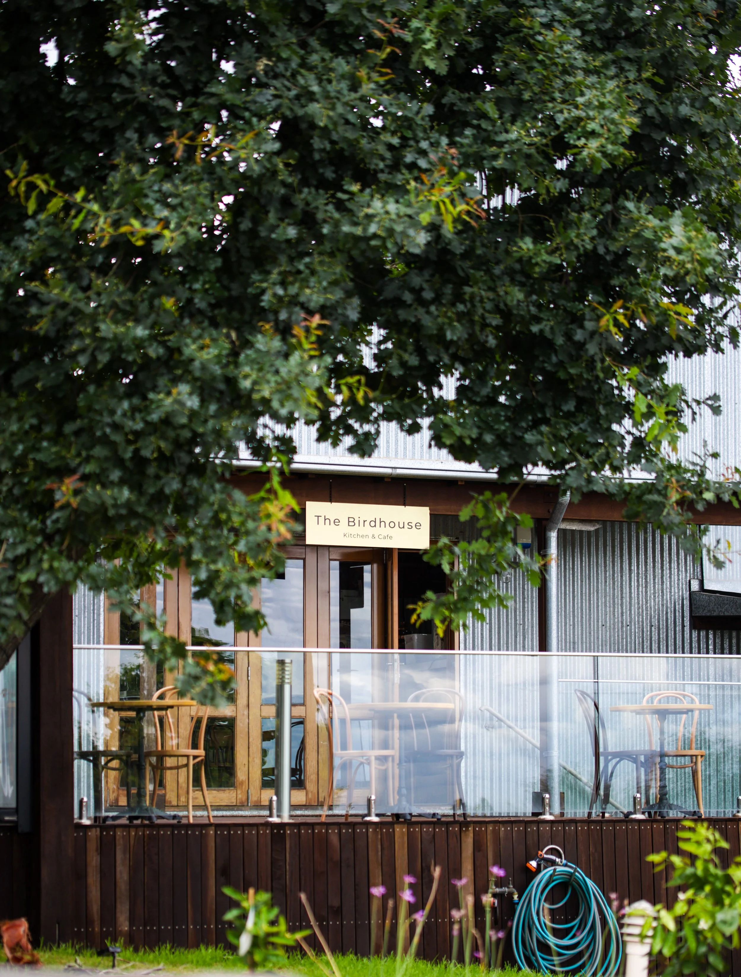 Outdoor view of The Birdhouse Kitchen & Cafe with wooden patio furniture outside, surrounded by greenery, and a hose coiled near the fence.