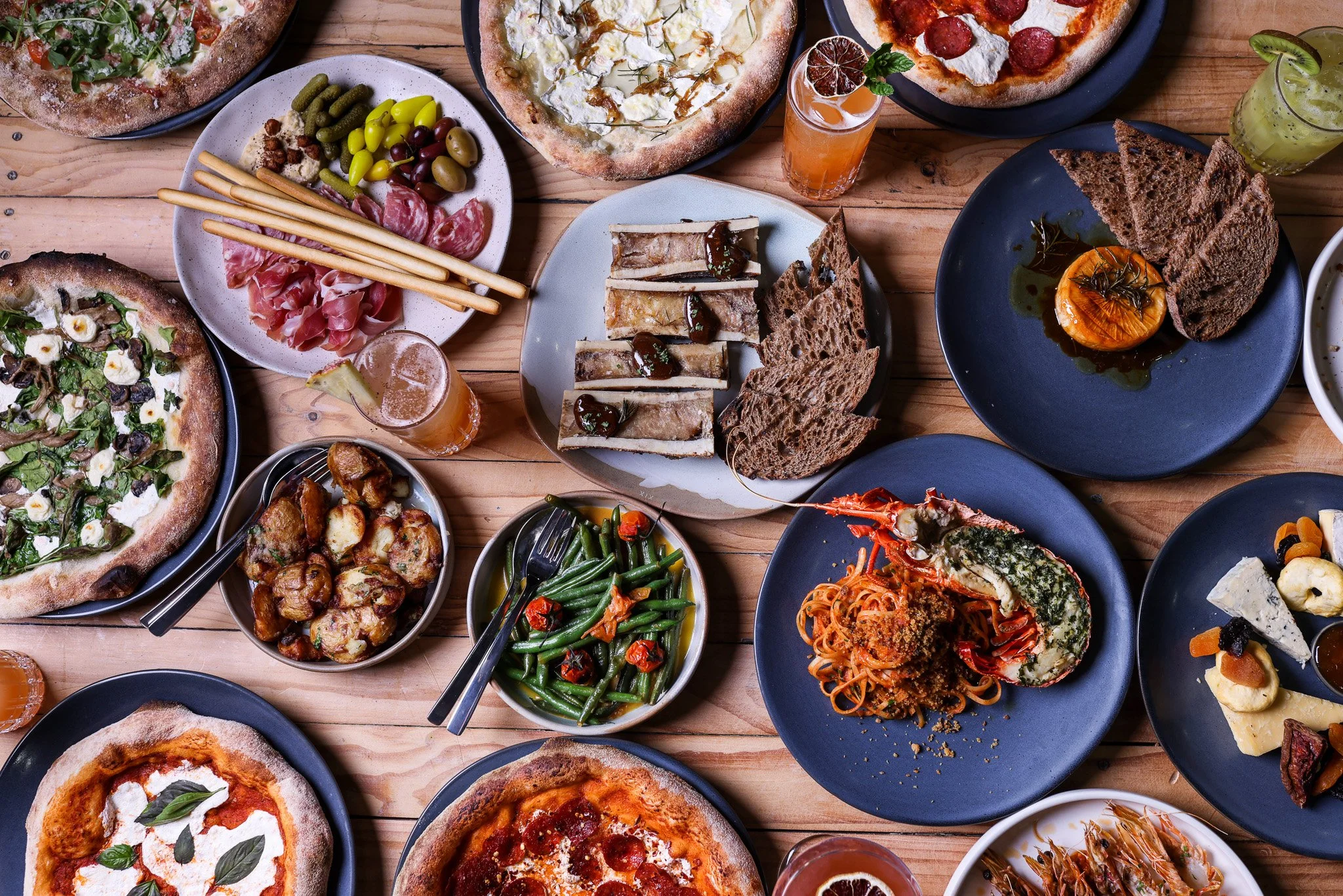 A spread of various dishes on a wooden table, including pizza, salads, bread, charcuterie, pasta, grilled vegetables, and drinks.