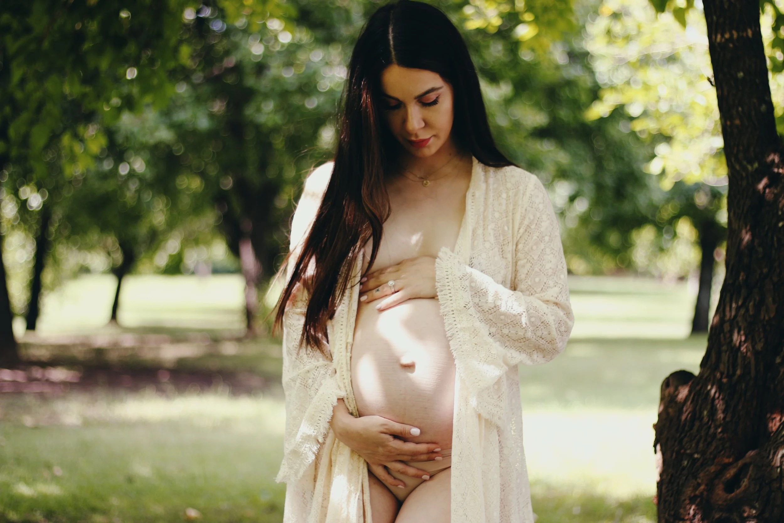 Pregnant woman in a cream lace robe standing outdoors among trees, gently holding her belly with one hand and touching her chest with the other, in a lush green park setting.