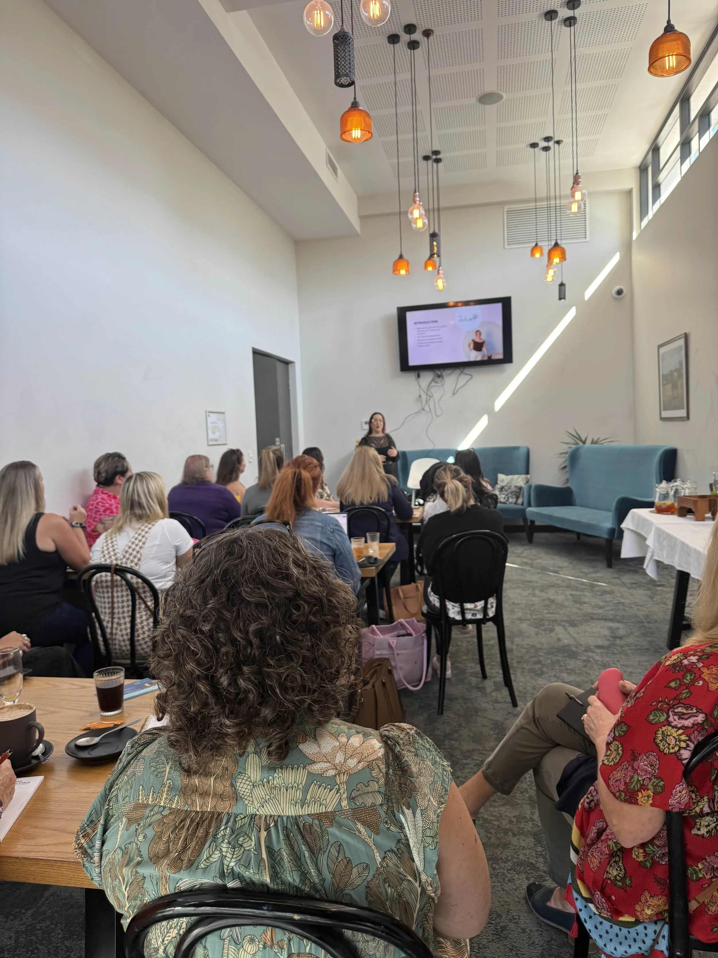 Women attending a presentation in a modern conference room with hanging pendant lights, a large screen, and blue seating.