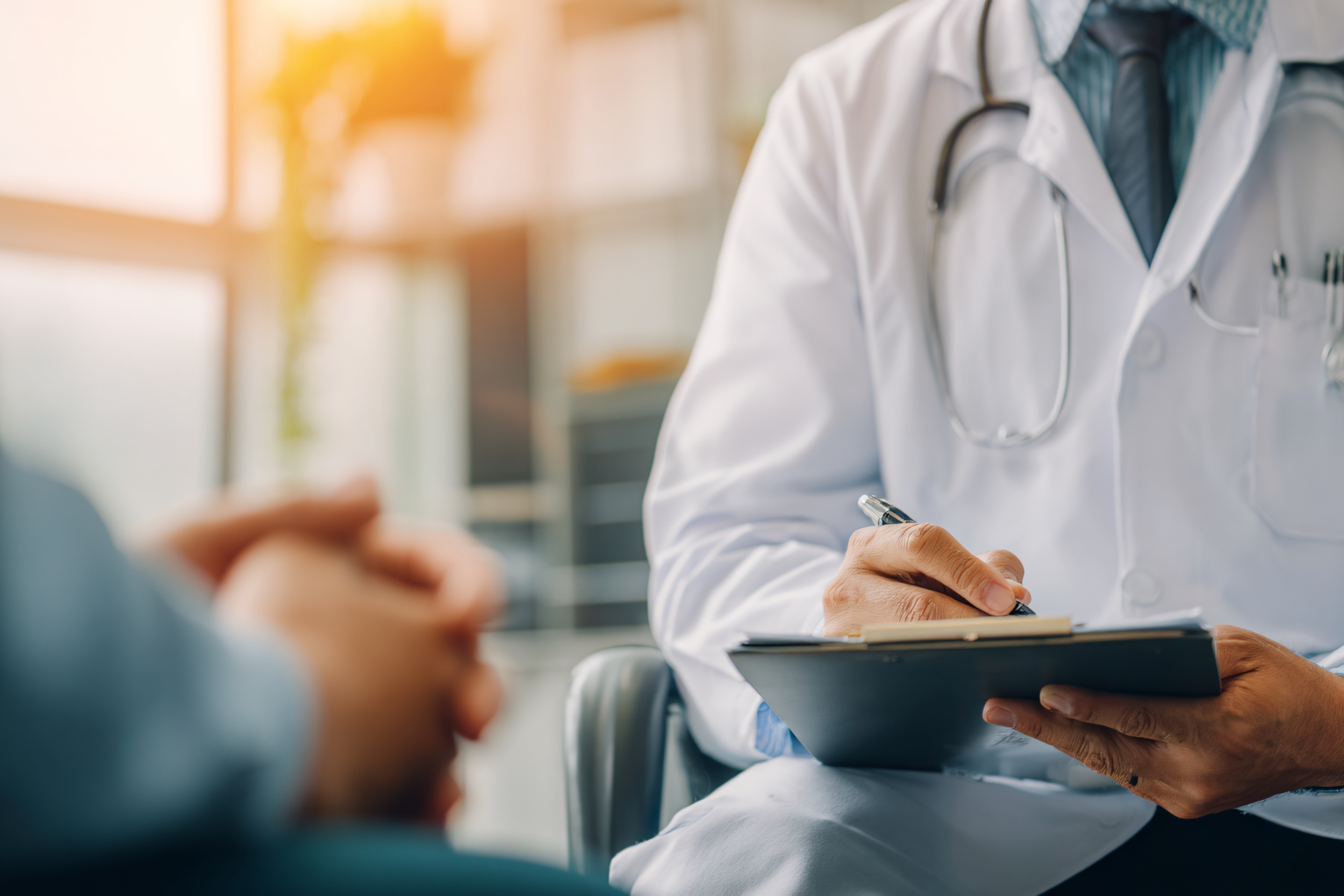 A doctor in a white coat with a stethoscope around his neck writing notes on a clipboard during a medical consultation.