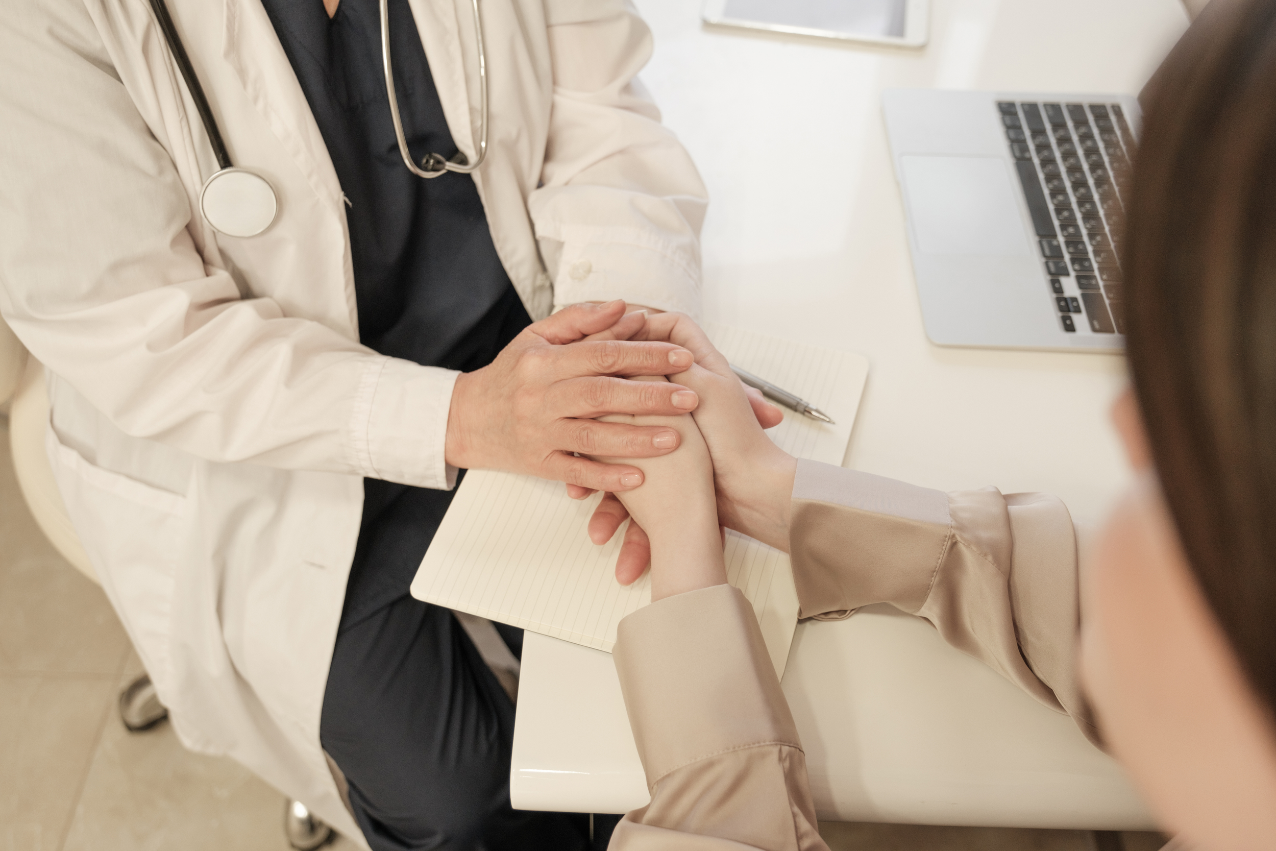 Medical professional holding a patient's hand during consultation with a stethoscope around their neck, sitting at a white table with a notepad and laptop.