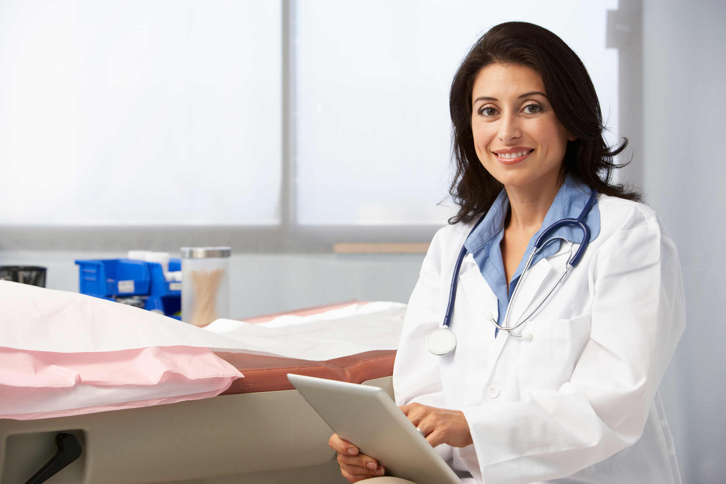A female doctor in a white coat smiling, holding a tablet, in a medical examination room with an examination table and medical supplies in the background.