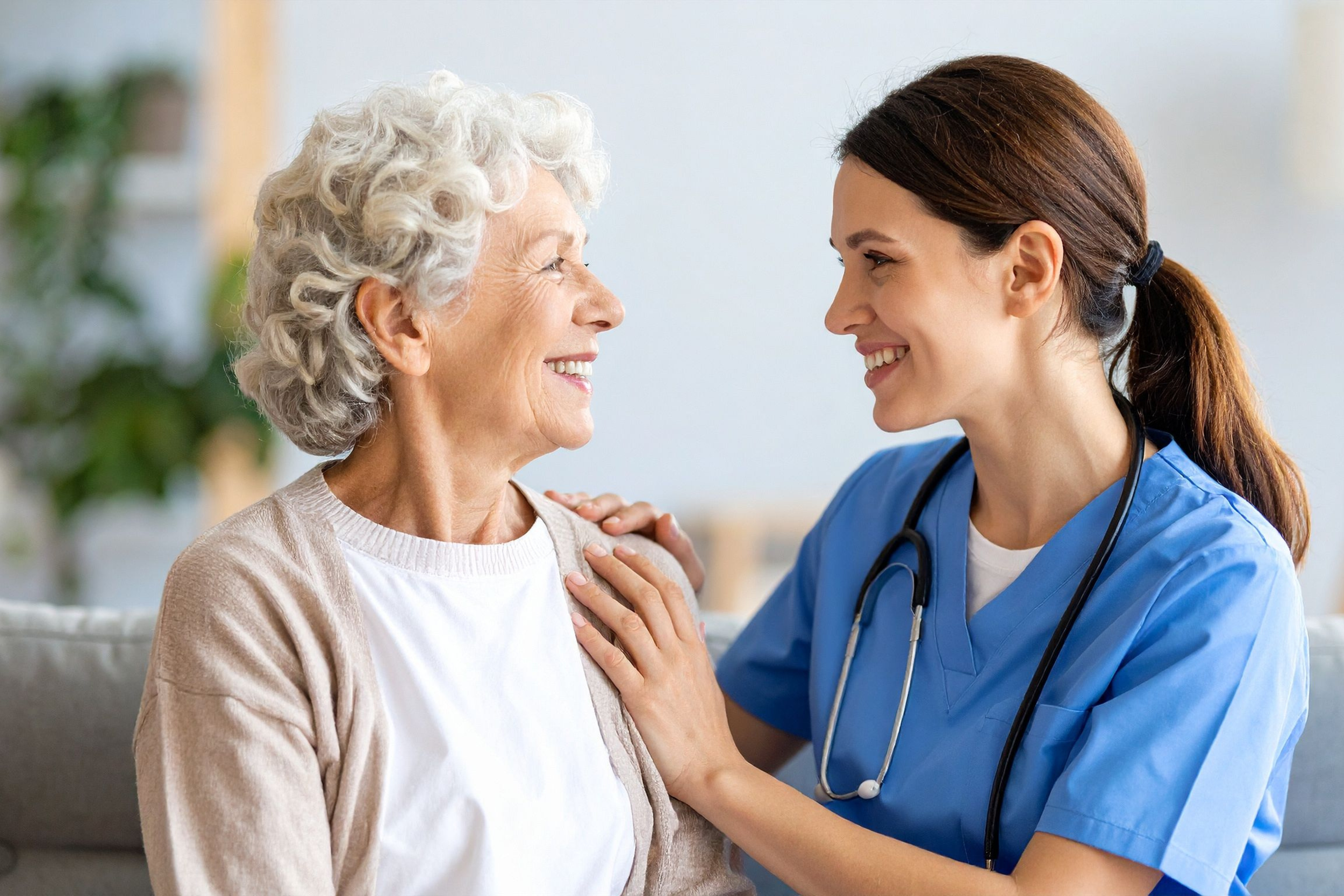 A nurse in blue scrubs and a stethoscope smiling and holding an elderly woman's shoulder, both smiling and making eye contact in a medical setting.
