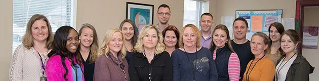 Group of diverse adults standing together in a classroom or office, smiling at the camera.