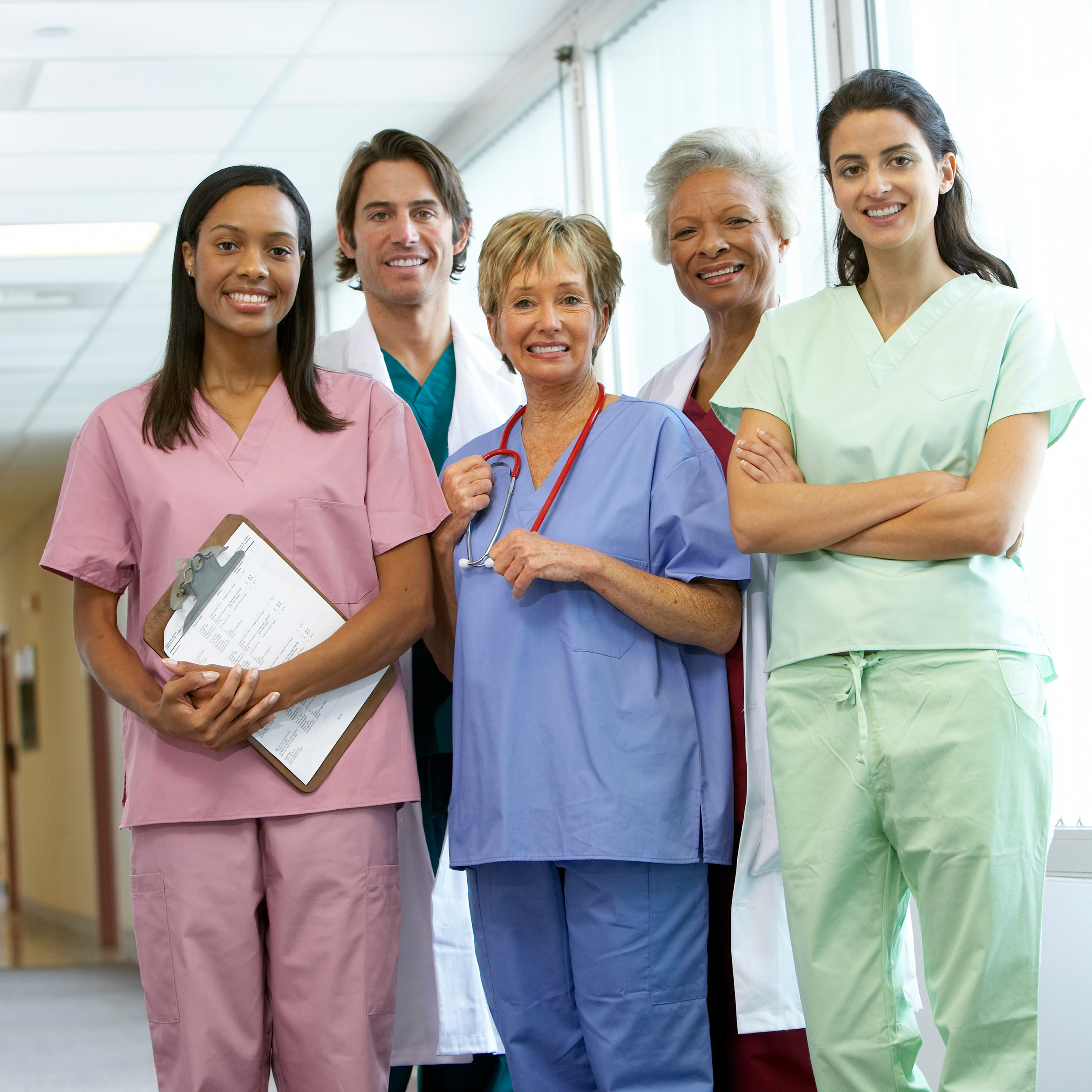 Group of five diverse healthcare professionals in scrubs and lab coat standing together in hospital corridor, smiling.