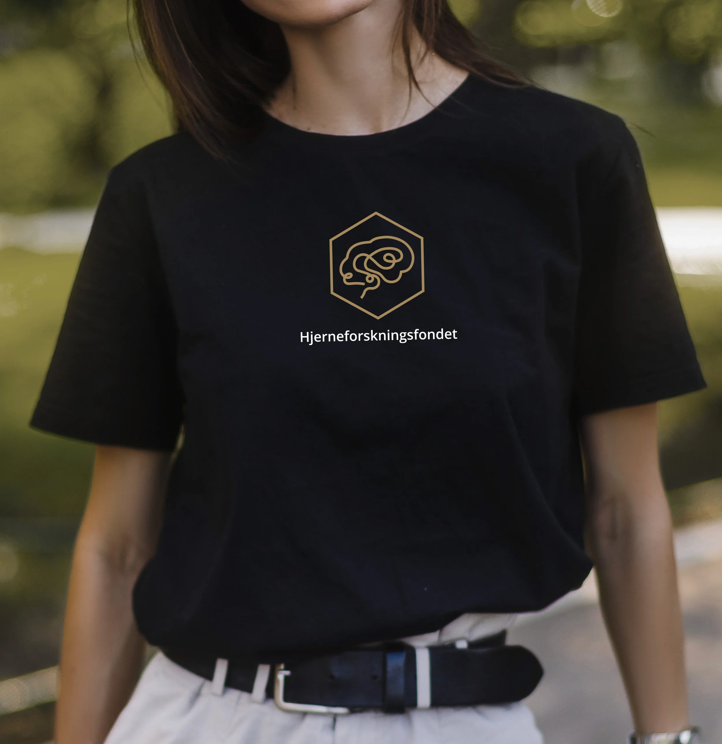 Person som bærer svart t-skjorte med logo og teksten 'Hjerneforskningsfondet' på brystet, ute i naturen.