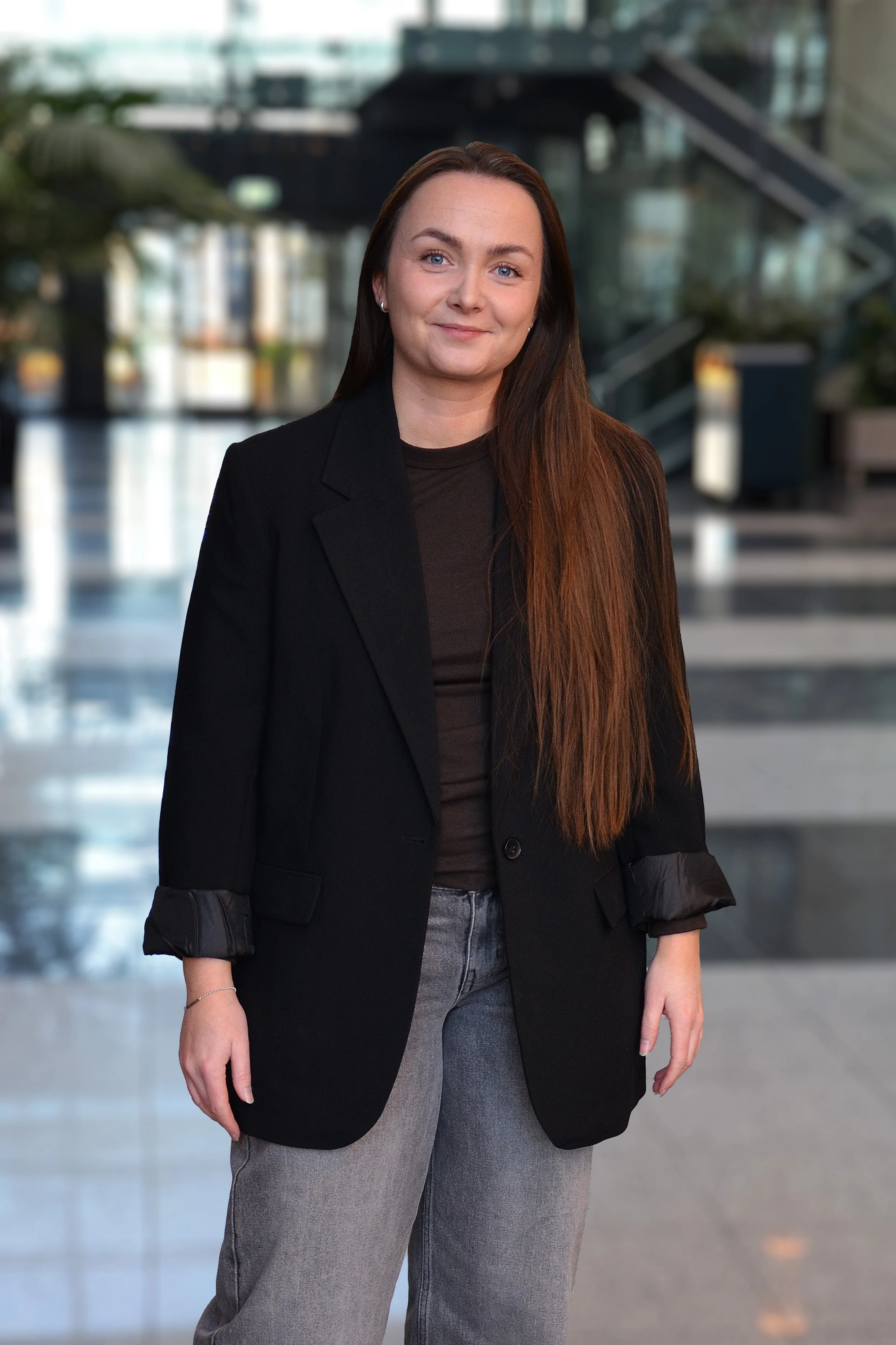 En kvinne med langt brunt hår, iført svart blazer og svarte bukser, står i en moderne innendørs bygning med store vinduer og trapp i bakgrunnen.