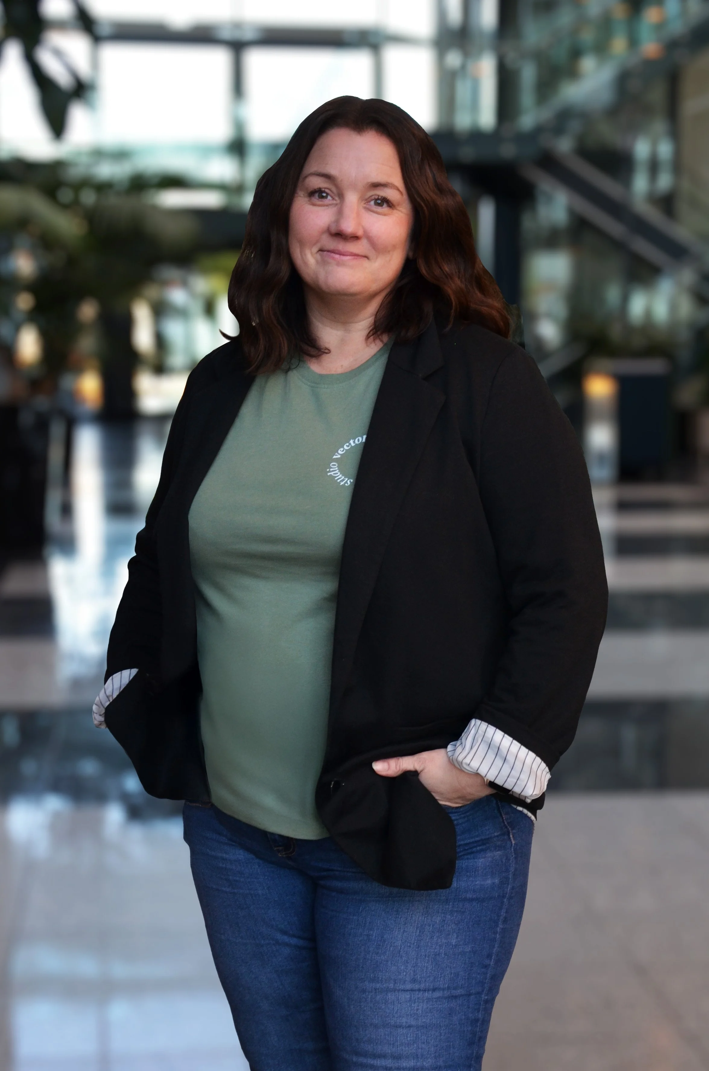 En kvinne med brunt, bølget hår smilende i en moderne innendørs atrium med glassvegger og trapper i bakgrunnen.