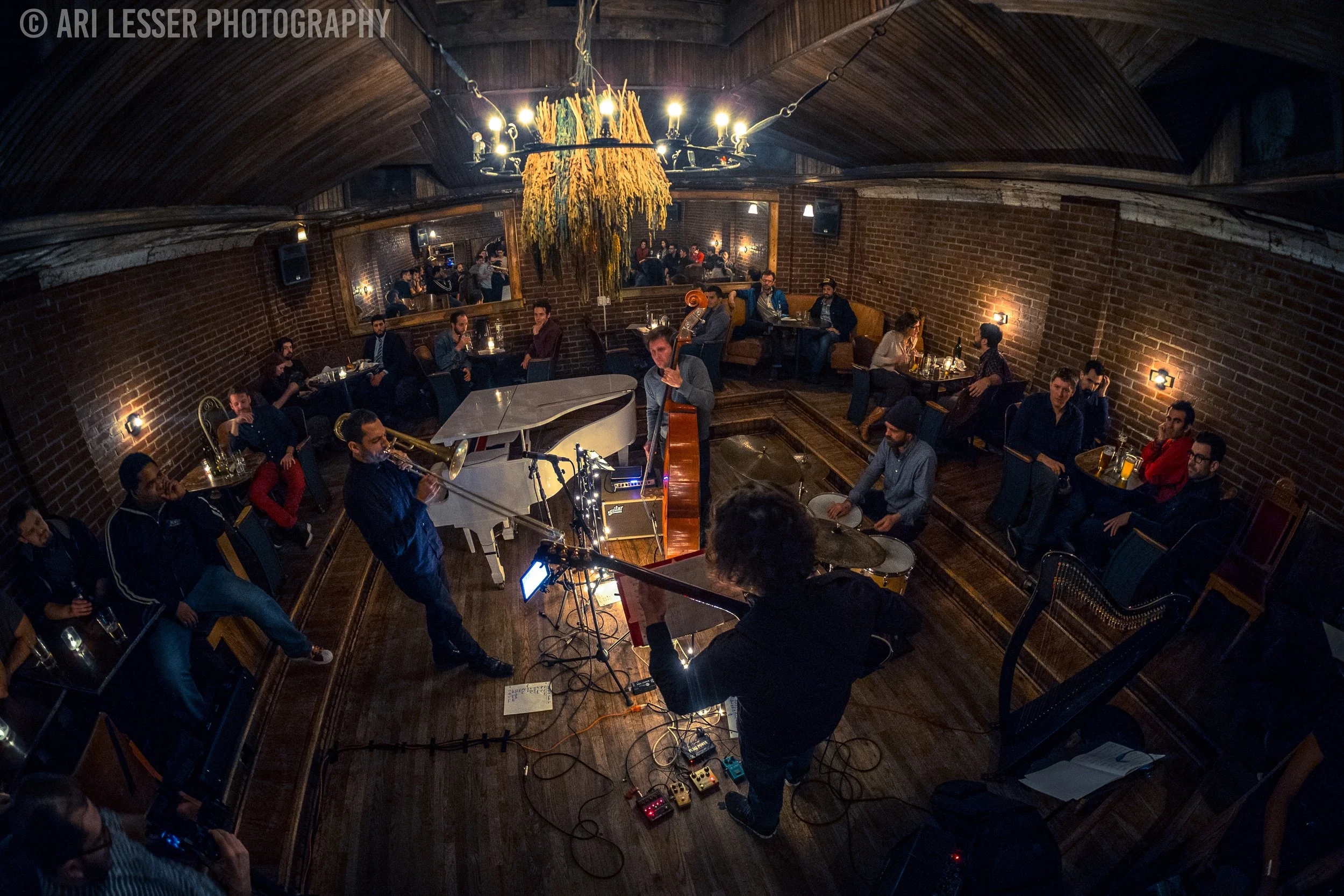 Jazz band performing live music in intimate venue with brick walls, wooden ceiling, and small tables with audience members watching, some with drinks, and a large mirror reflecting the scene.