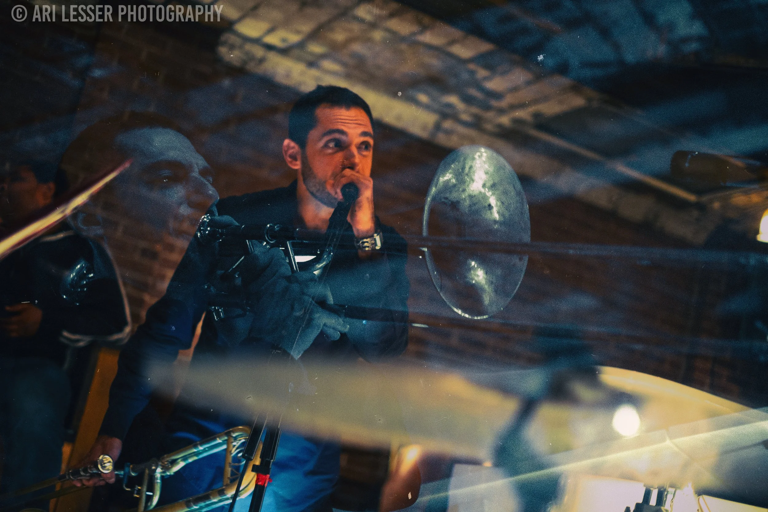 Musicians performing live on stage, with one playing the saxophone and the other singing into a microphone, inside a venue with brick walls and wooden ceiling.