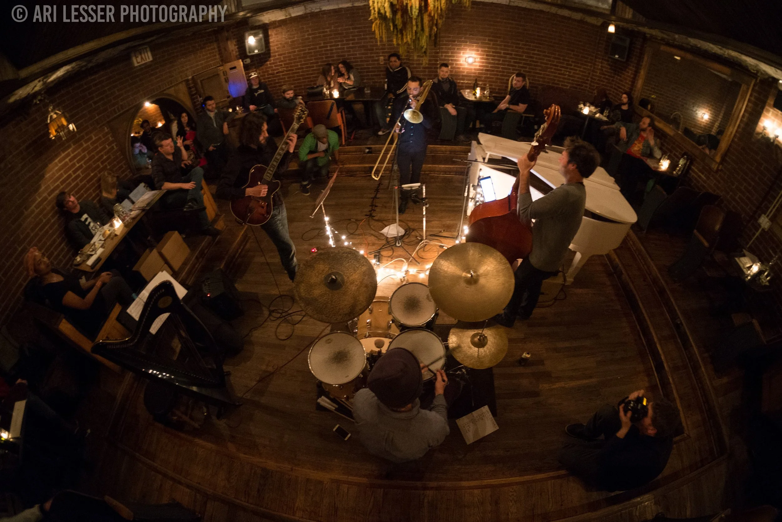 A band performs live music in a cozy, dimly lit venue with brick walls. Musicians include a guitarist, trombone player, upright bassist, and drummer. Audience members sit around tables, some enjoying drinks, while one person takes photographs.
