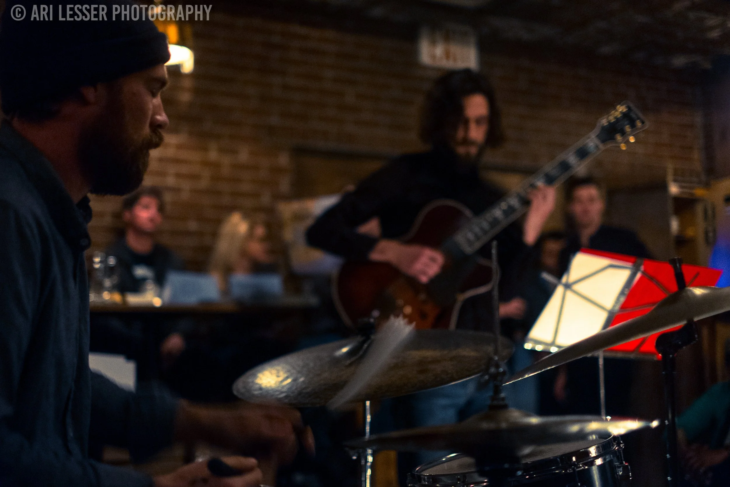 Musicians performing at a dimly lit live music venue, with a drummer in the foreground playing a drum set and a guitarist in the background playing an acoustic guitar.