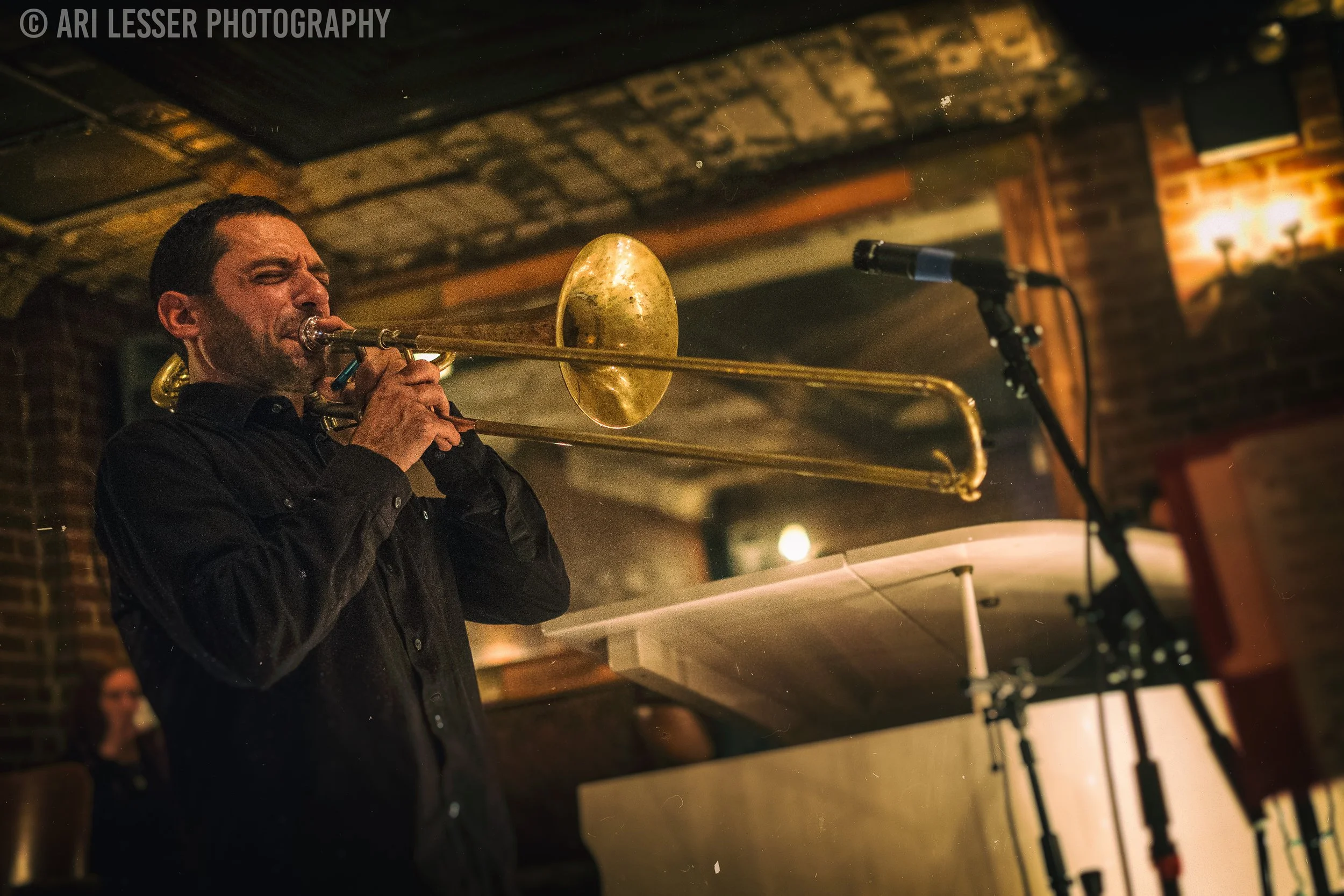 A man playing a trombone, with closed eyes, in a dimly lit room with brick walls, microphone stand, and a woman in the background.