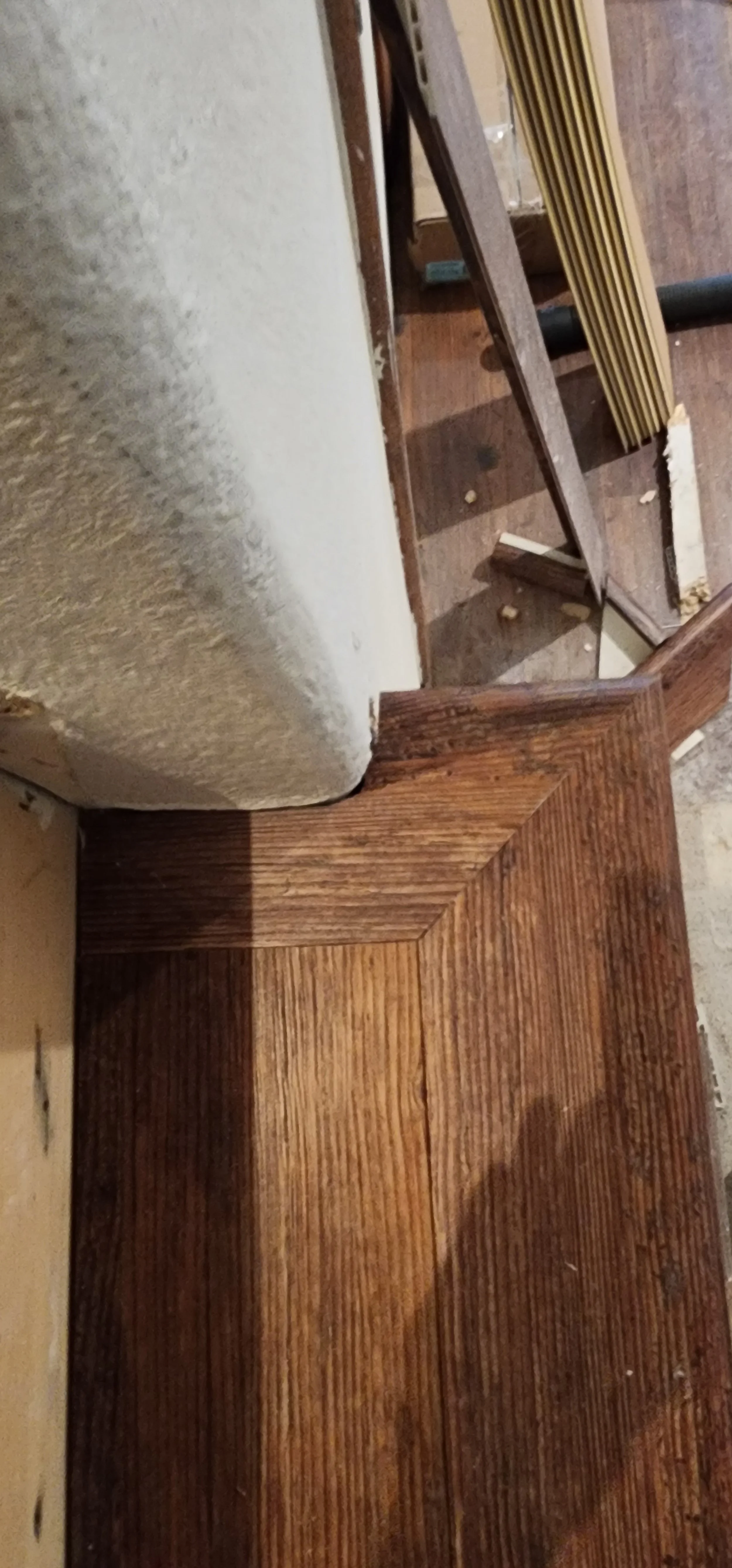 Close-up of a partially finished wooden piece of furniture or flooring with a rich, dark finish, next to a white wall with visible signs of wear or sanding.