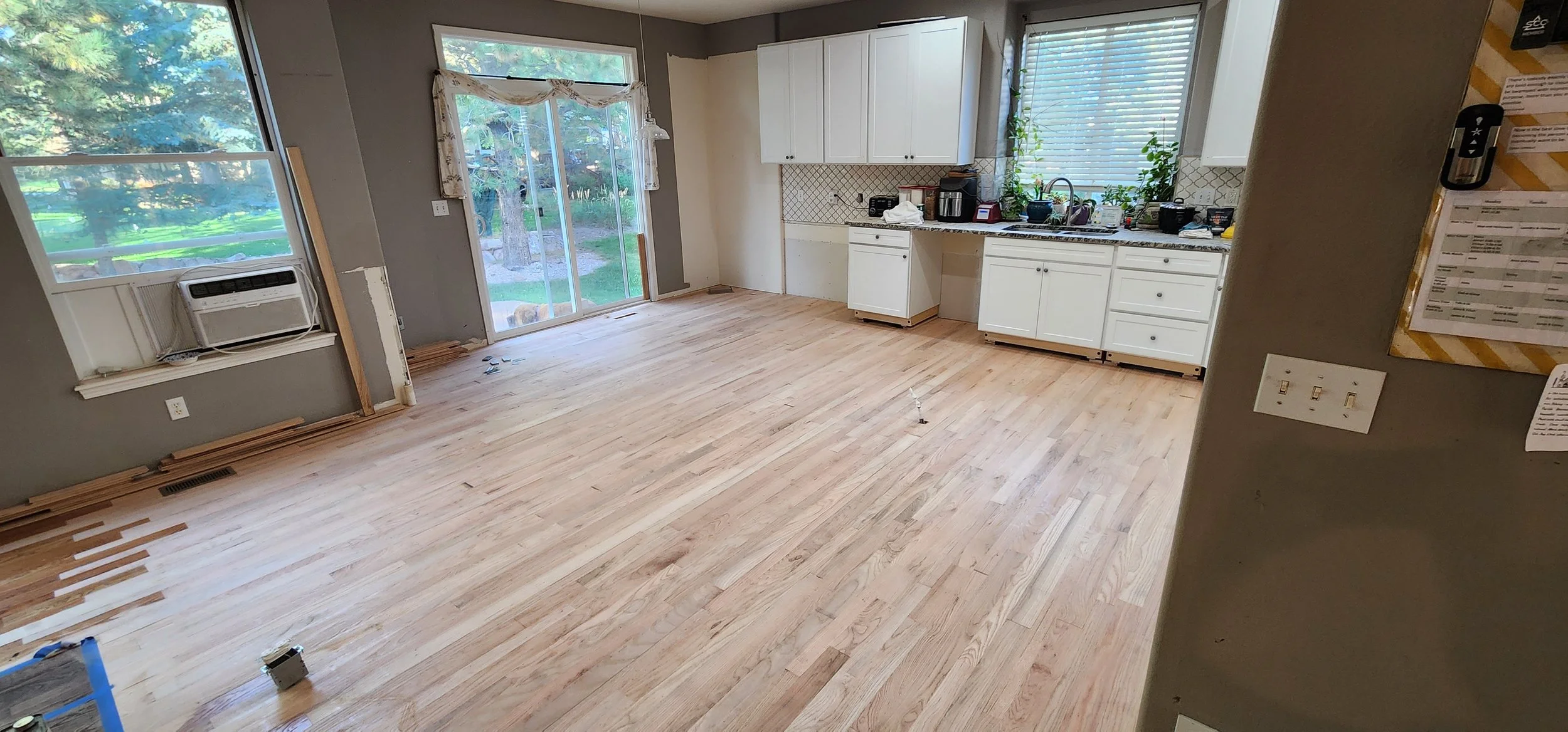 Empty kitchen and dining area with hardwood floors, white cabinets, and large windows showing greenery outside.