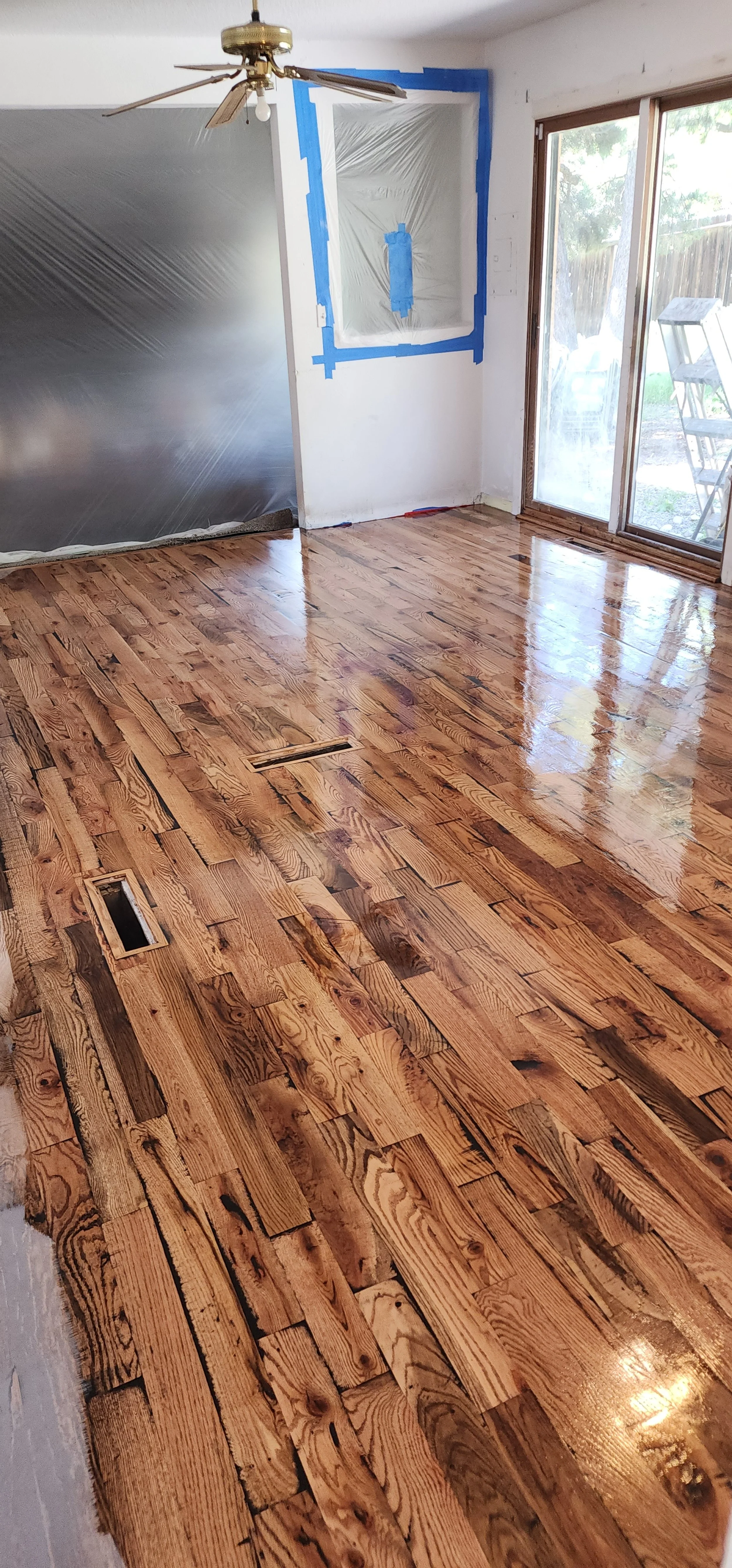 A hardwood floor is being installed in a bright room with sliding glass doors. The floor appears freshly sanded and polished. Part of the wall is covered with plastic sheeting, and there is blue painter's tape around a window opening. A ceiling fan i