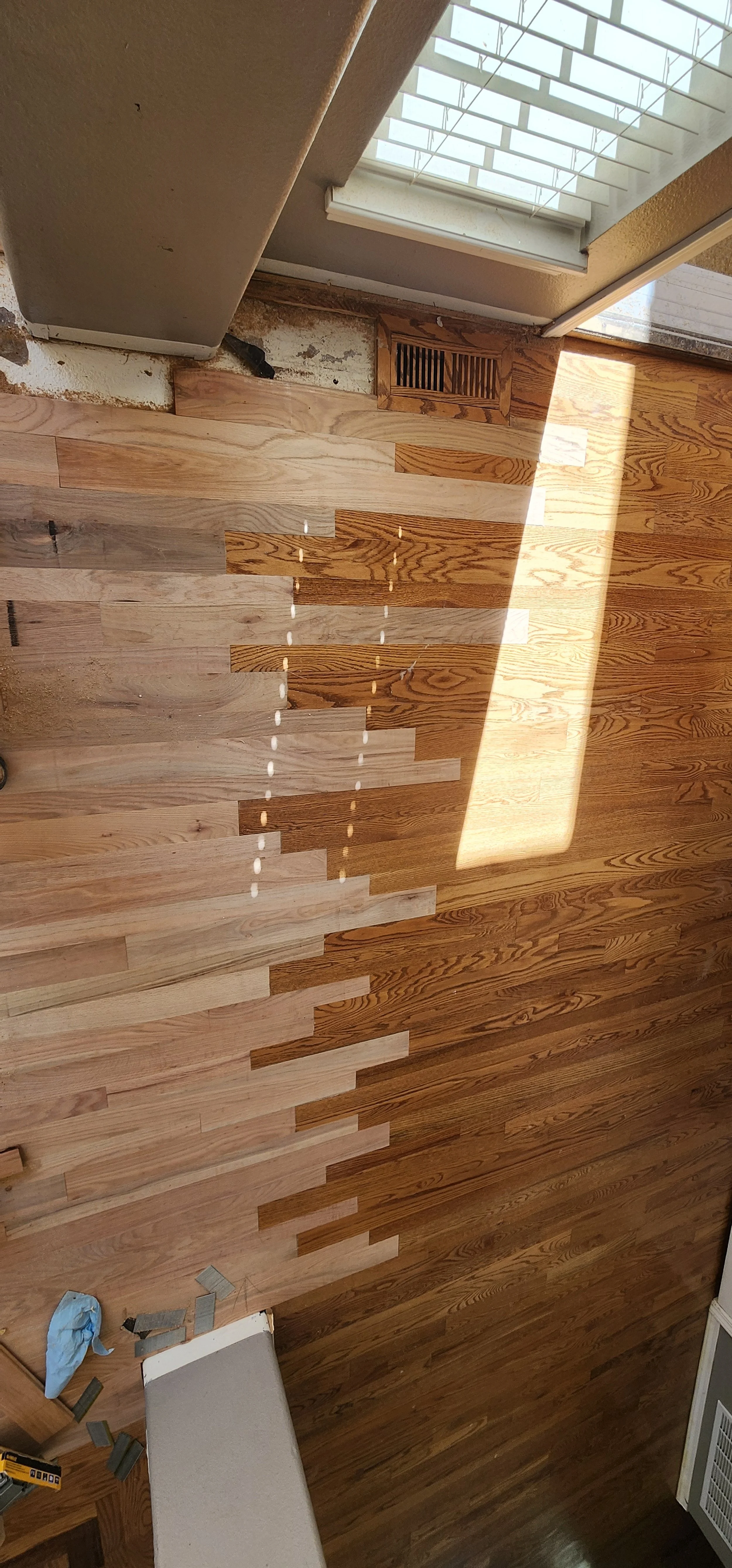 A hardwood floor under construction, with some planks not yet installed. Sunlight streams in through a skylight, casting light and shadow across the floor. Part of a vent, window, and some tools are visible.