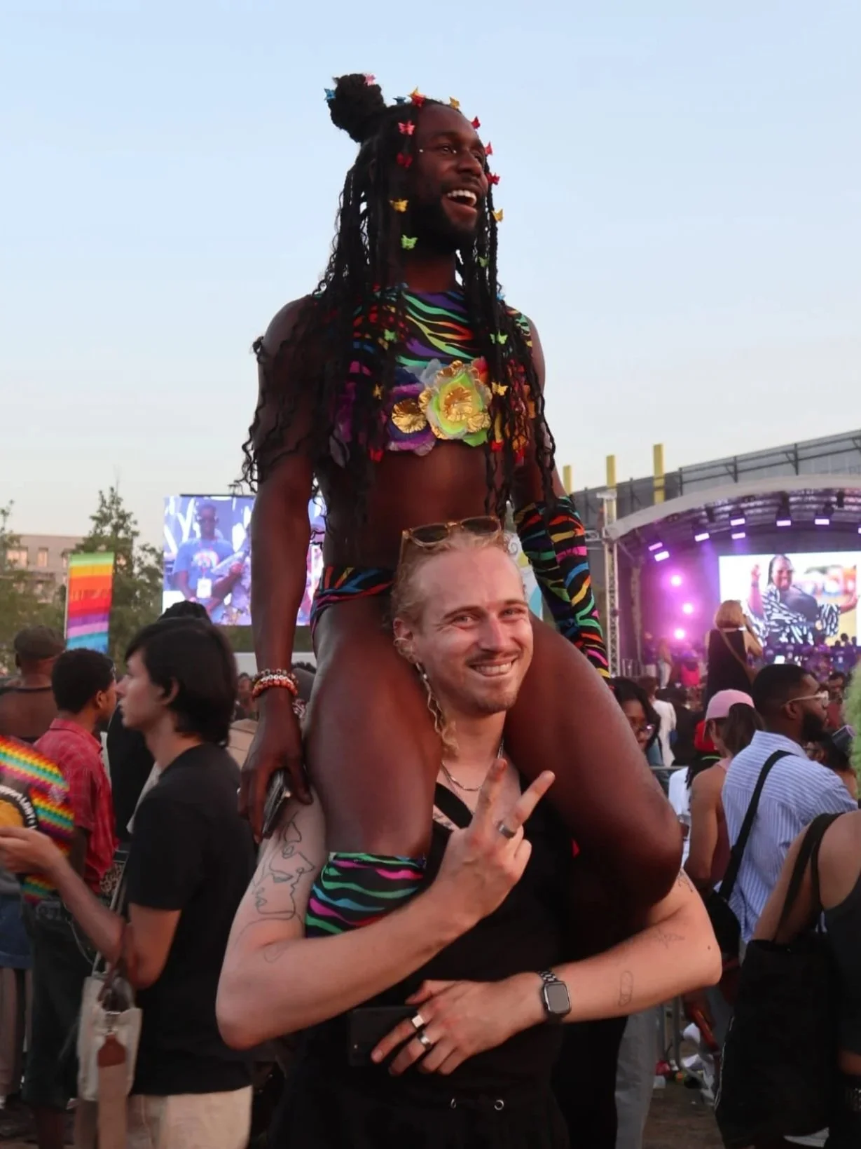 A man smiling and making a peace sign with his hand in front of a crowd at an outdoor concert, carrying a man on his shoulders; the man is wearing a colorful, striped outfit and has braids with butterflies in h hair, and the background shows a stage with purple lighting.