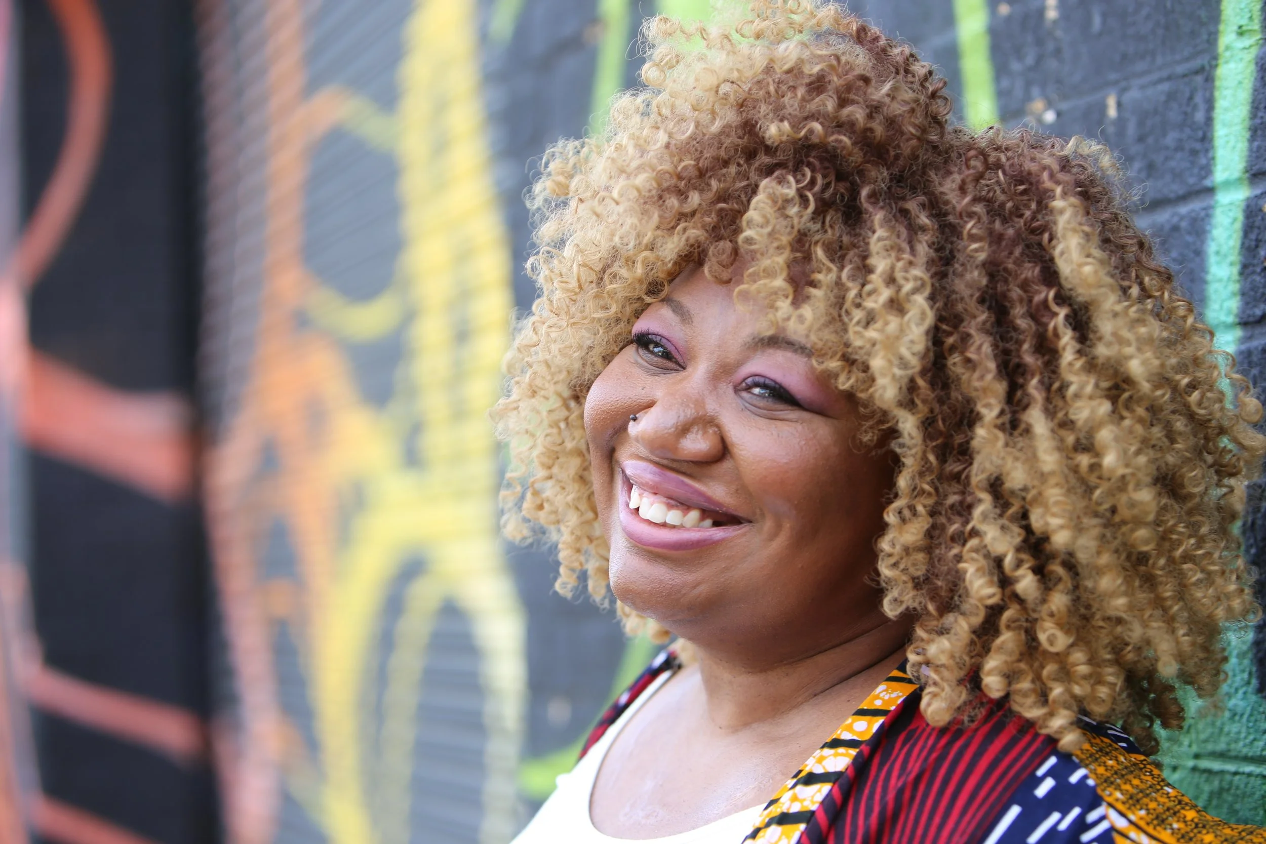 Charmaine Jean-Paul, founder and CEO of Rebel Led Equality, EDI consultant in Peckham, London. Close-up of a woman with curly blonde hair smiling against a black brick wall with colourful graffiti.