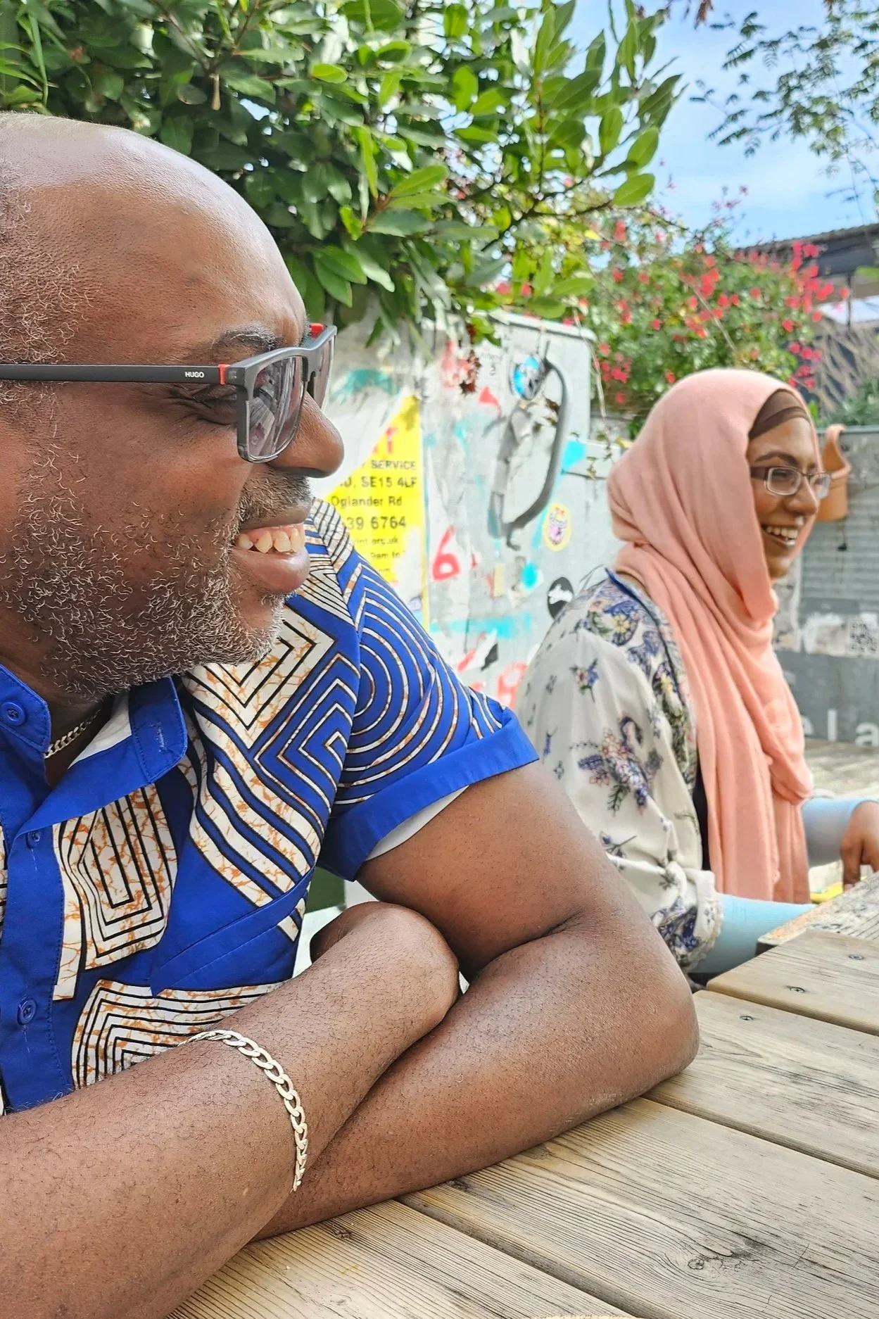 Two laughing people sitting outdoors at a wooden table, with green foliage and pink flowers in the background. The man in the foreground has glasses, a beard, and is wearing a blue patterned shirt. One woman has a pink headscarf and glasses.