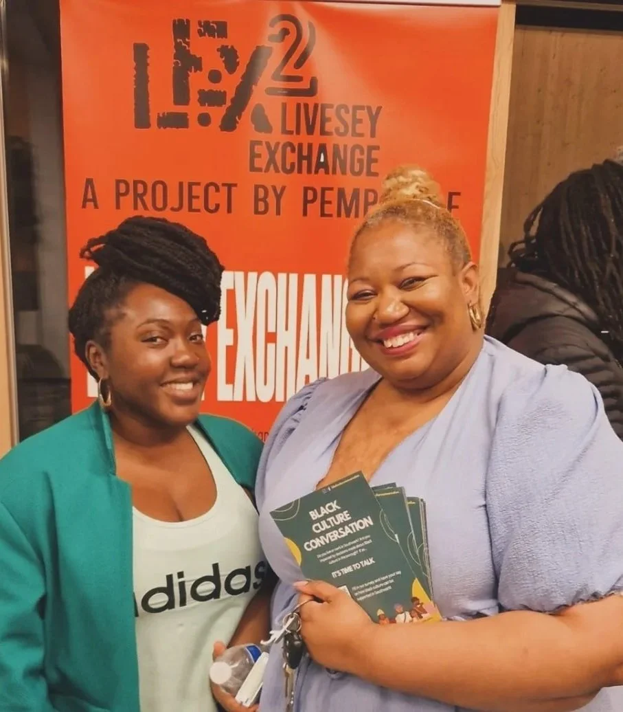 Two women smiling at a community event, holding flyers titled 'Black Culture Conversation', with an orange sign in the background that reads '12.2 Livesey Exchange, A Project by Pemba'.