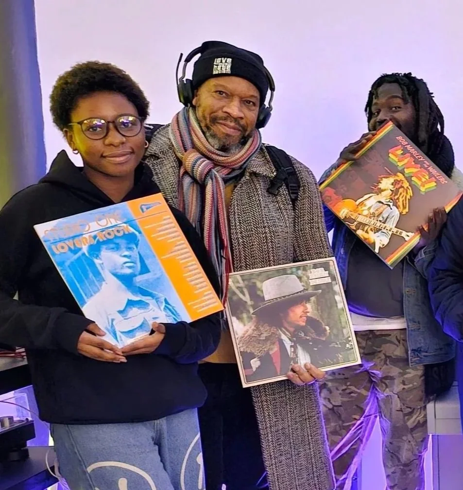 Three people standing indoors, holding vinyl records, with a purple-lit background