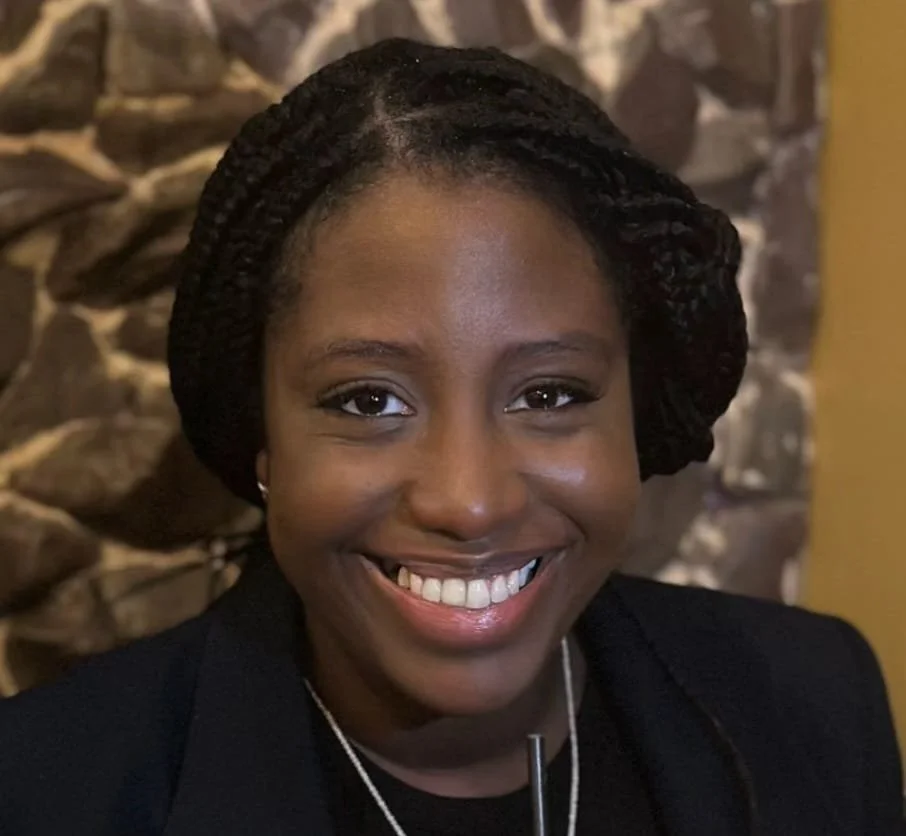 Close-up portrait of an African American woman smiling, wearing a black blazer, silver necklace, and earrings, with a stone wall background.