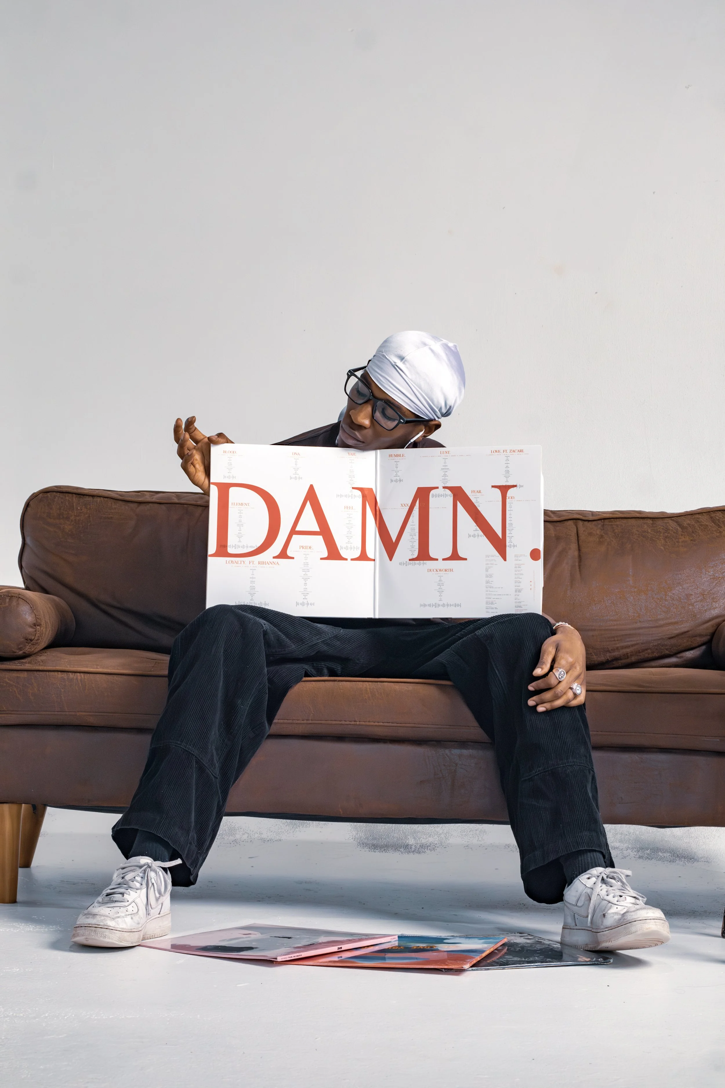 Person sitting on a brown sofa, wearing glasses, a white head wrap, and a ring, holding a large album cover with the word ‘DAMN.’ in red letters.
