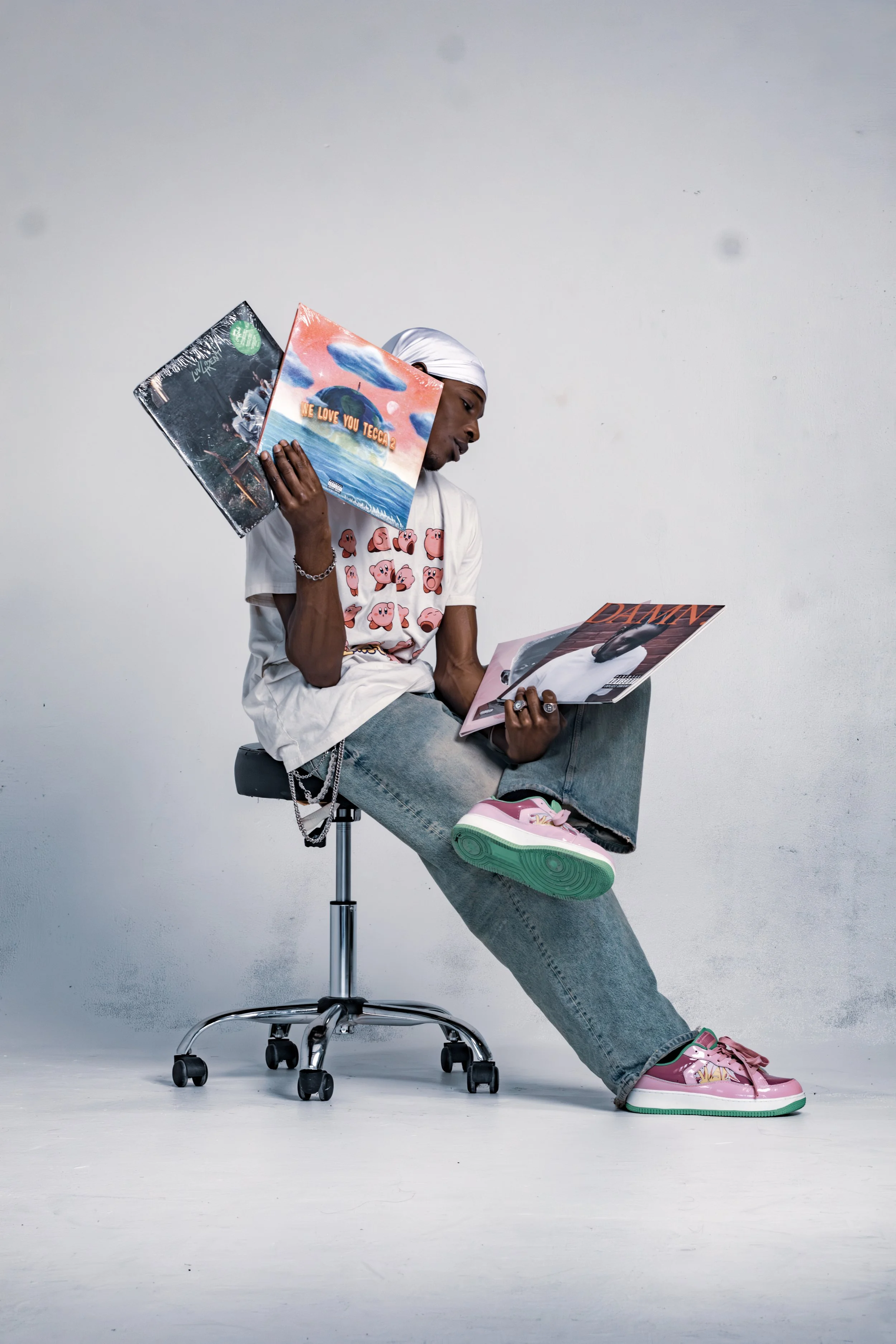 Person sitting on an office chair, wearing a white t-shirt with cartoon characters, grey jeans, and pink sneakers, holding vinyl records and reading a magazine against a plain white background.