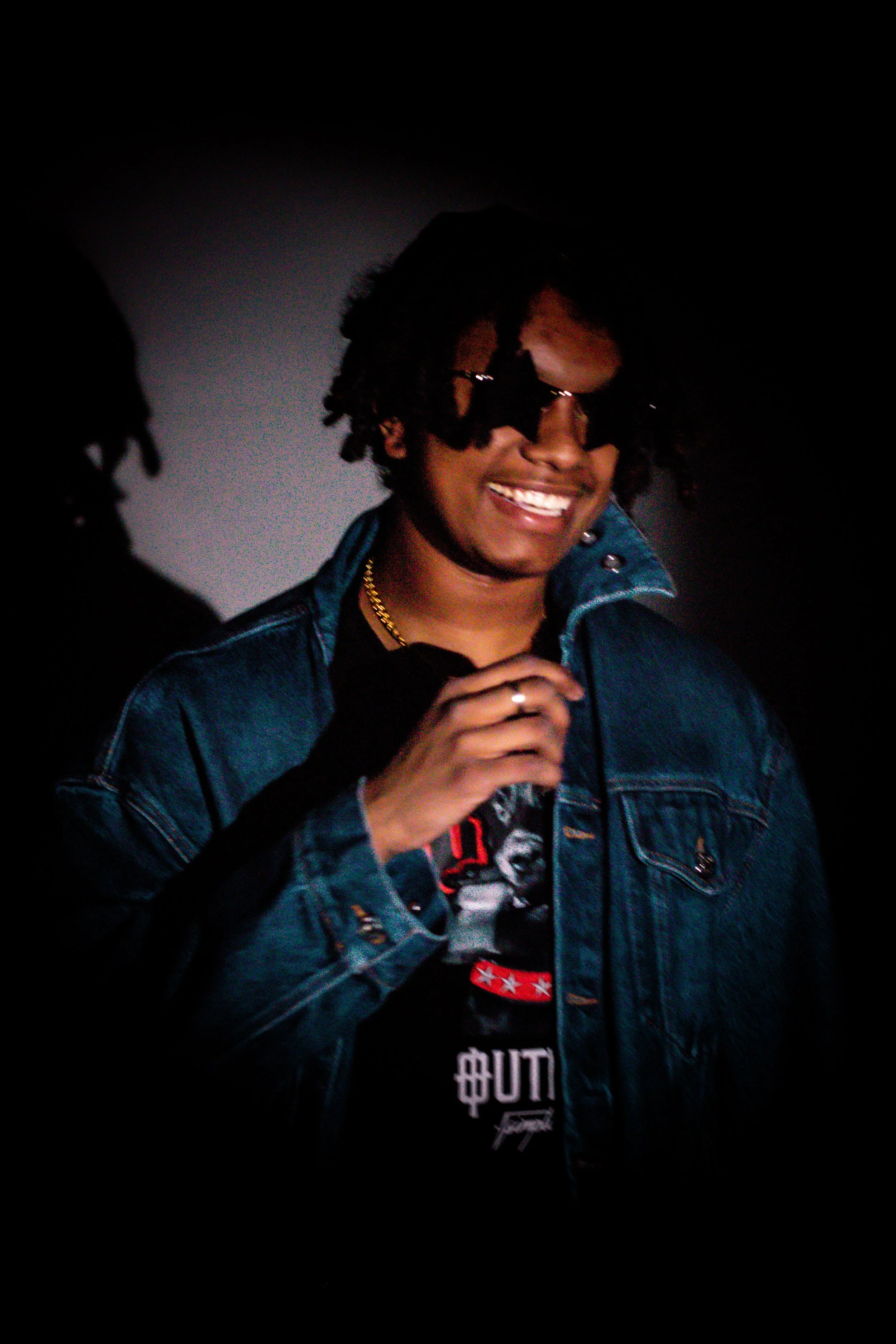 A young man with dreadlocks, sunglasses, gold chain, and a denim jacket smiling and posing against a dark background.