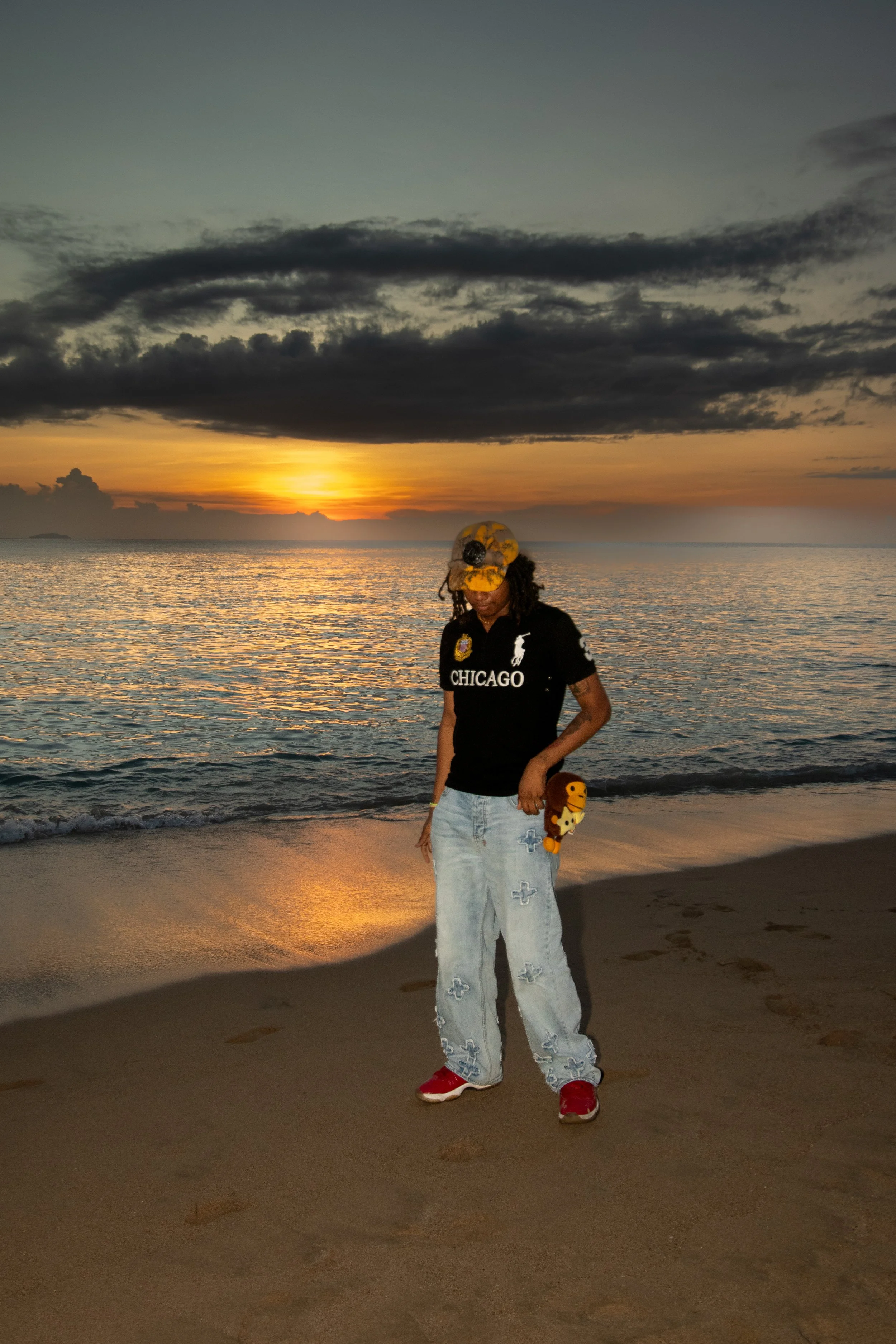 Person standing on a beach during sunset, wearing a black polo shirt with 'CHICAGO' printed on it, light blue ripped jeans, red sneakers, a camouflage cap, and holding a small plush toy.