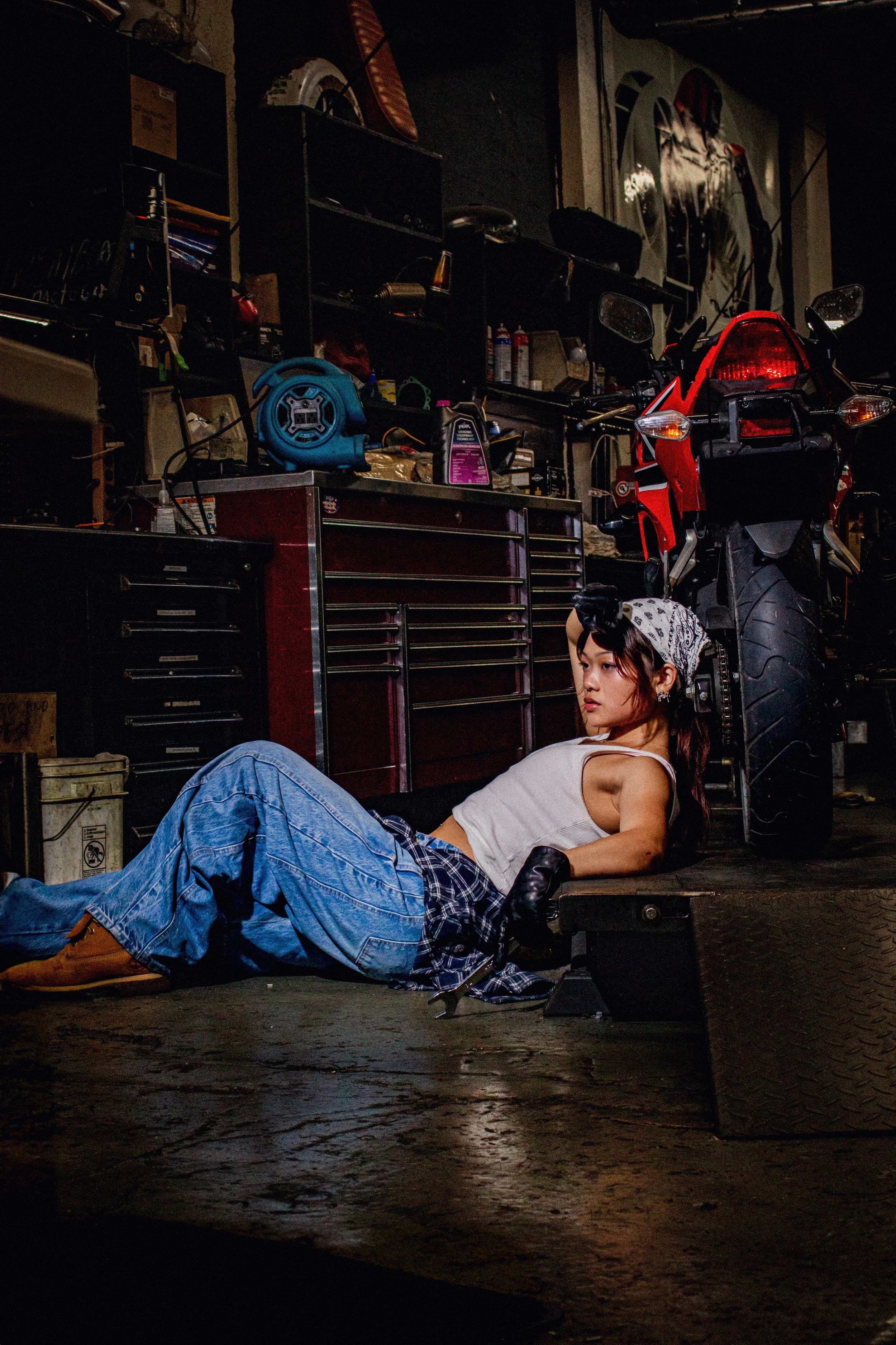 A young woman with a bandana and gloves lying on the floor of a garage, leaning on a bench next to a red motorcycle, with various tools and shelves in the background.