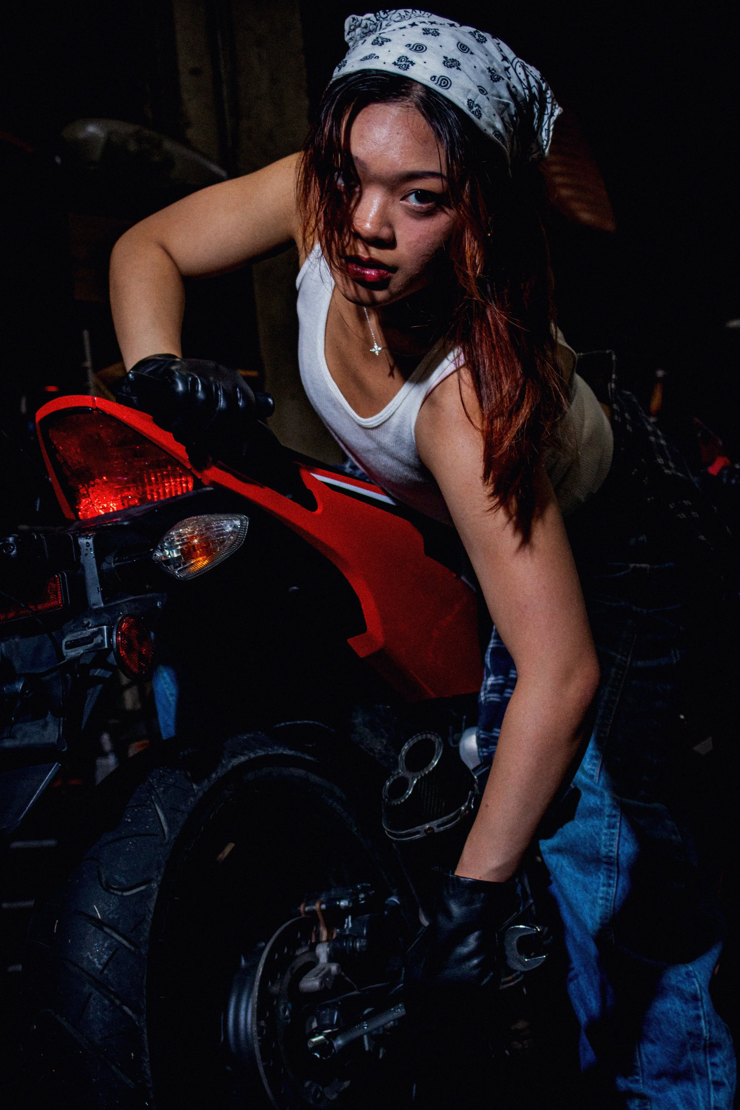 A woman with reddish hair, wearing a bandana, a white sleeveless top, and black gloves, leans over a red motorcycle in a dark indoor setting.