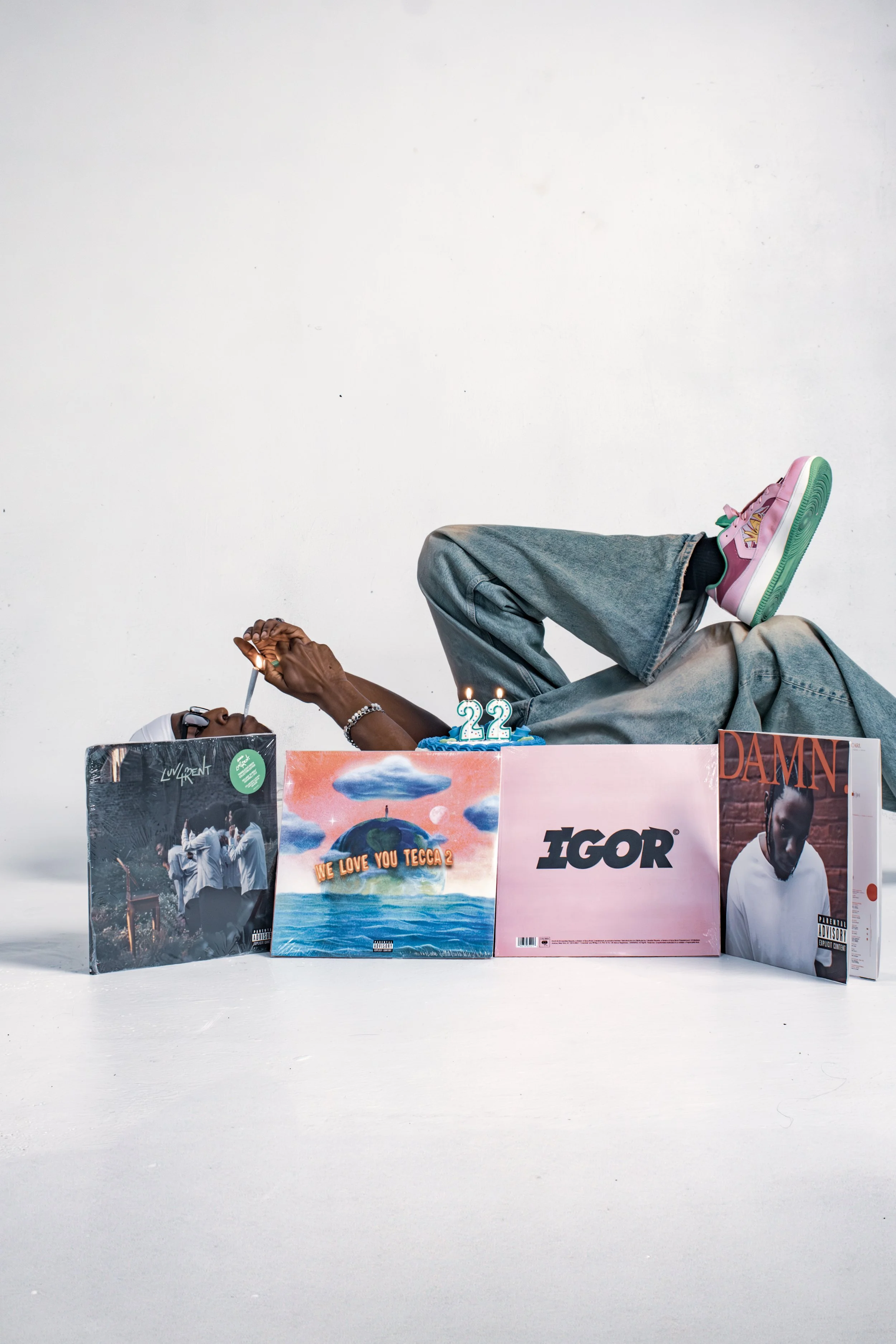 A person lying on their back on the floor, surrounded by four vinyl record albums, two candles, and a birthday cake with the number 22 candles on it. The person is wearing jeans, pink sneakers, and glasses, and is smoking.