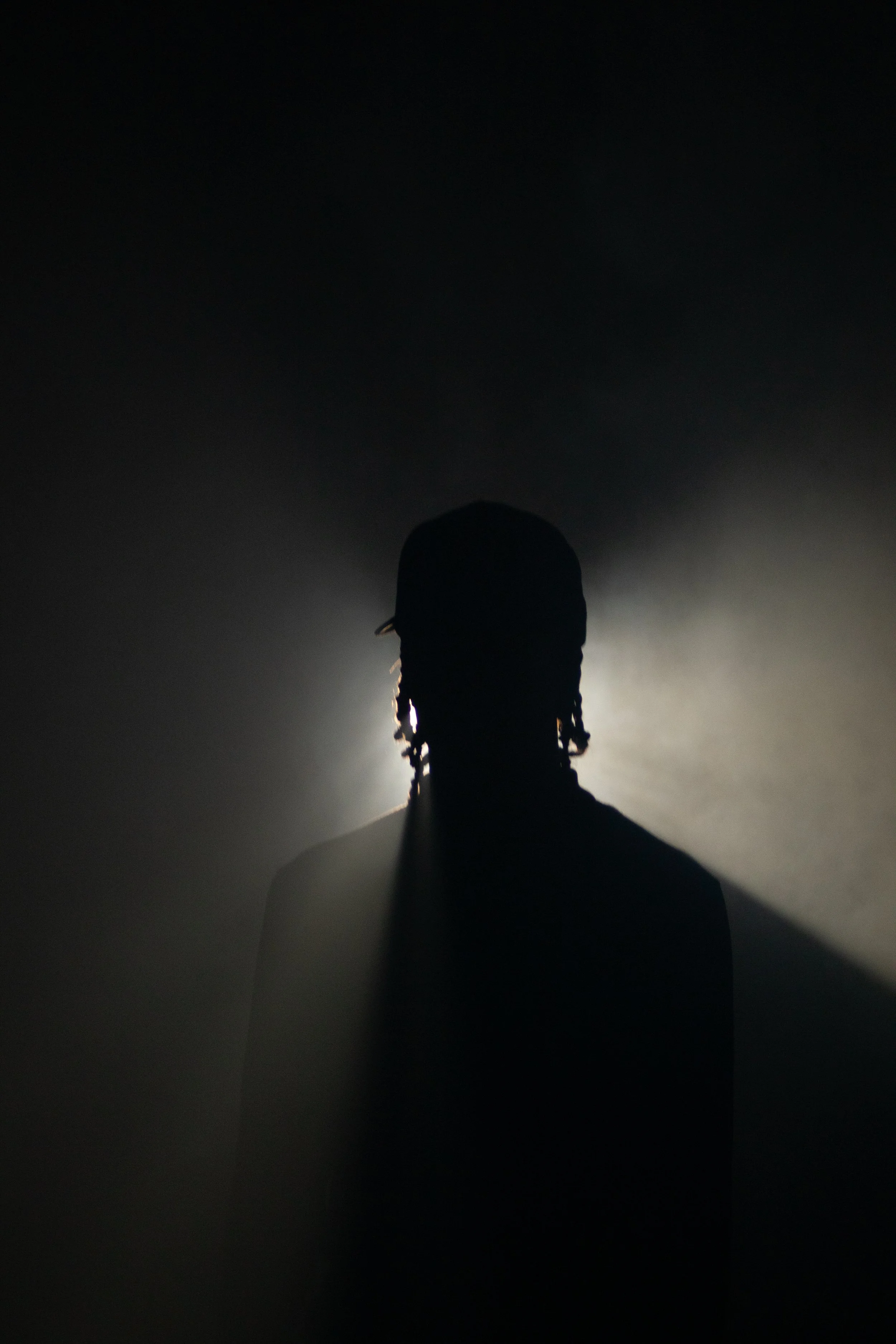 Silhouette of a person with dreadlocks facing backlit from behind, with a foggy or smoky background.