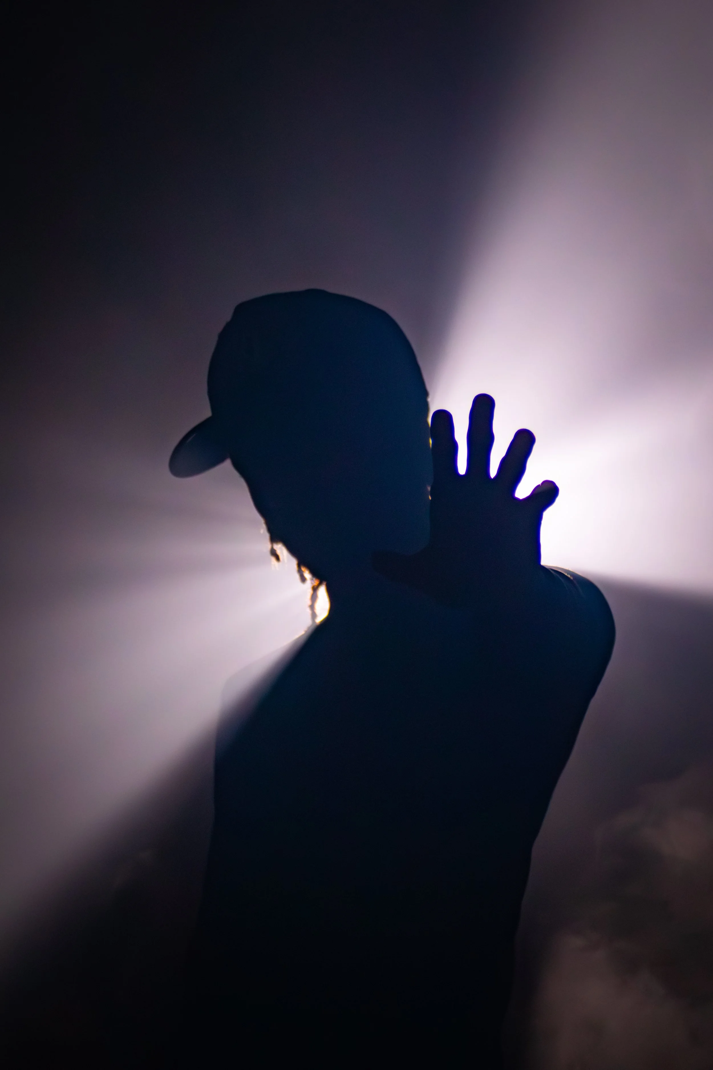 Silhouette of a person wearing a baseball cap, holding up their hand against a bright light background.