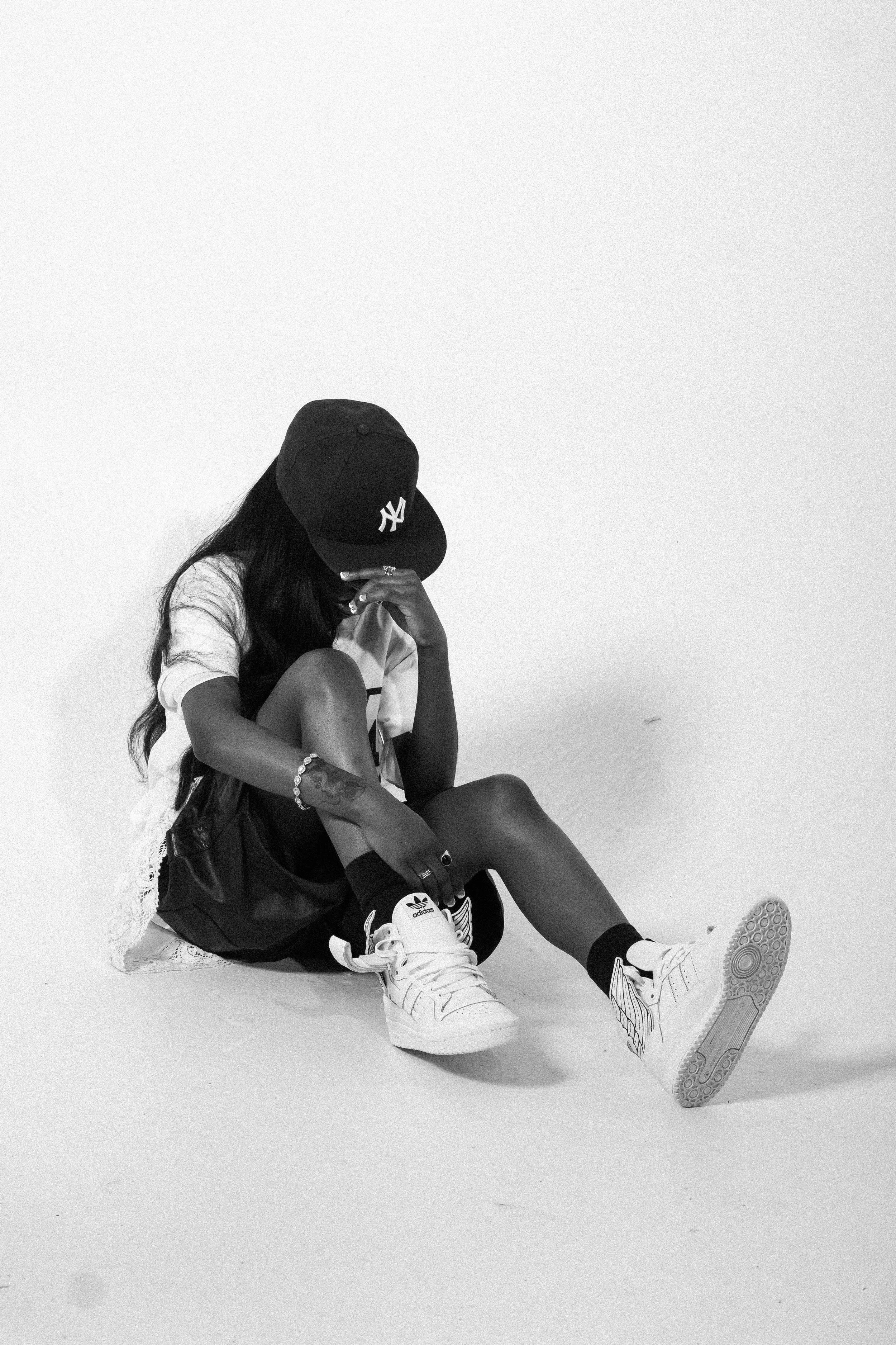 A young woman sitting on the floor, wearing a black baseball cap with the Yankees logo, a white t-shirt, black shorts, and white Adidas sneakers, with one hand resting on her knee and the other touching her face, against a plain white background.