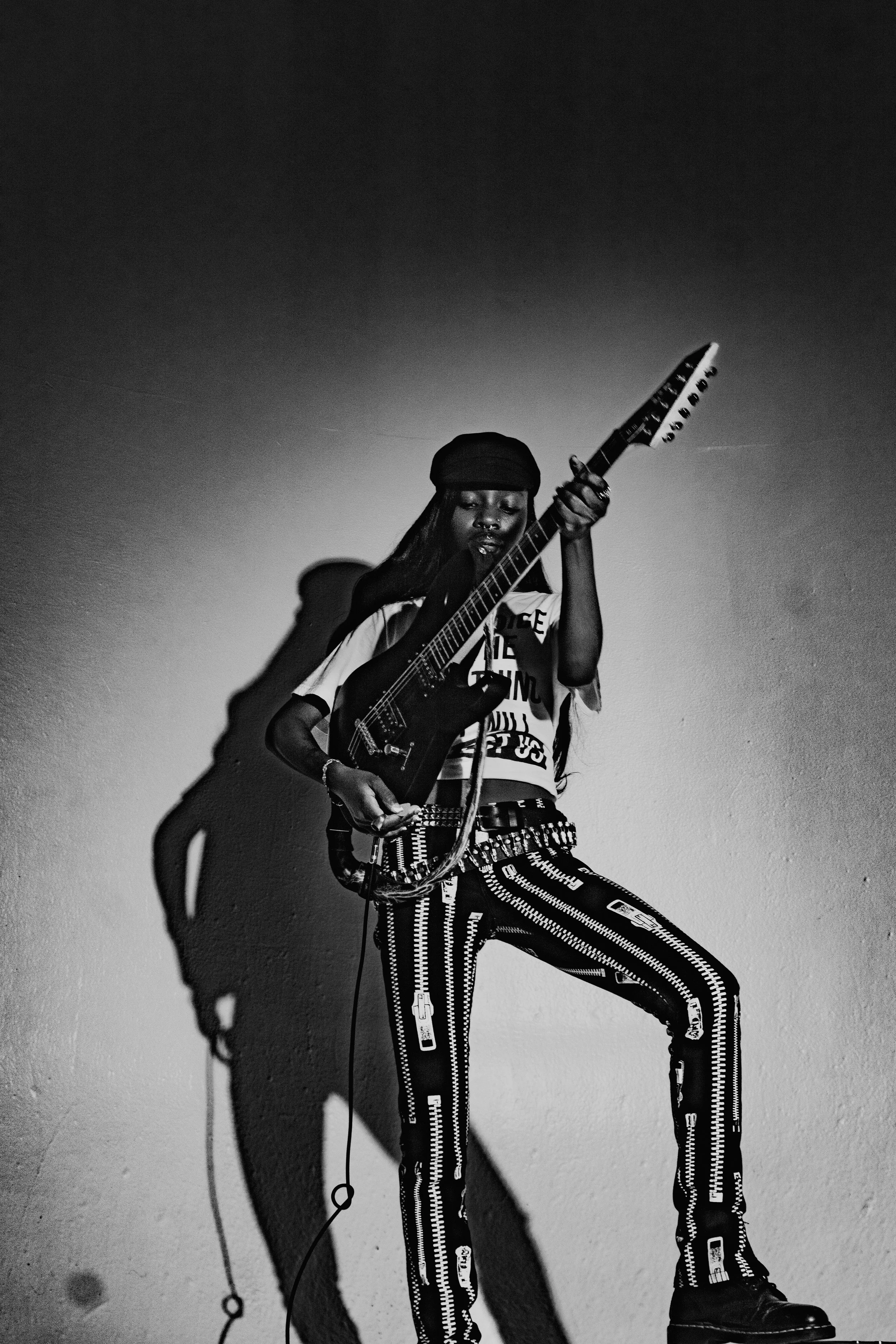 A woman playing an electric guitar, standing against a plain wall with a shadow cast on it. She is wearing a hat, a graphic T-shirt, and patterned pants.
