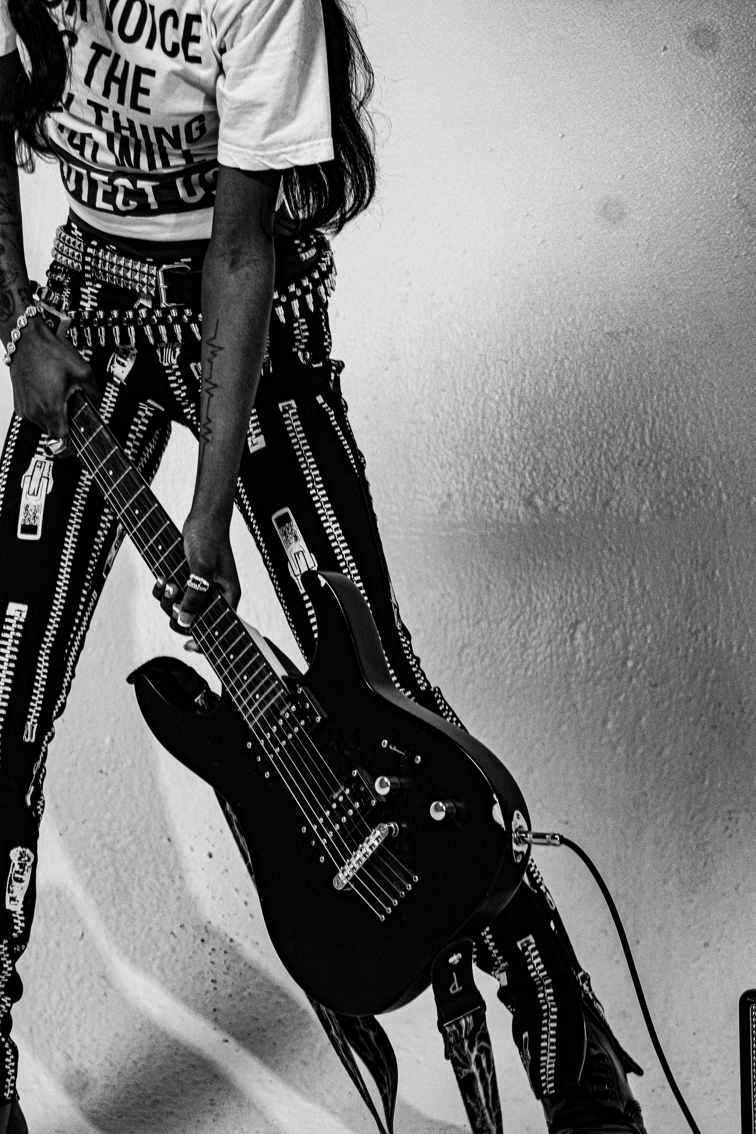 Person holding a black electric guitar, dressed in patterned pants, white T-shirt with bold text, and wearing several accessories, against a plain wall.