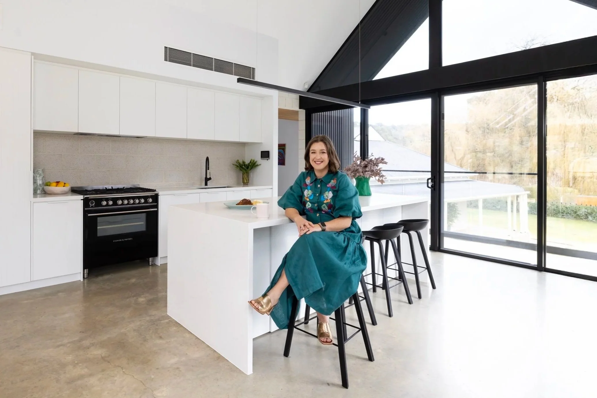 Bethany sitting on a black barstool at a white kitchen island in a modern, bright kitchen with large glass sliding doors.