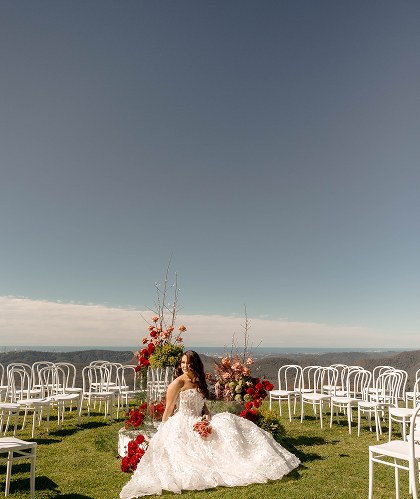 Bride in a white wedding gown sitting outdoors on green grass with flowers and chairs set up for a wedding ceremony under a blue sky.